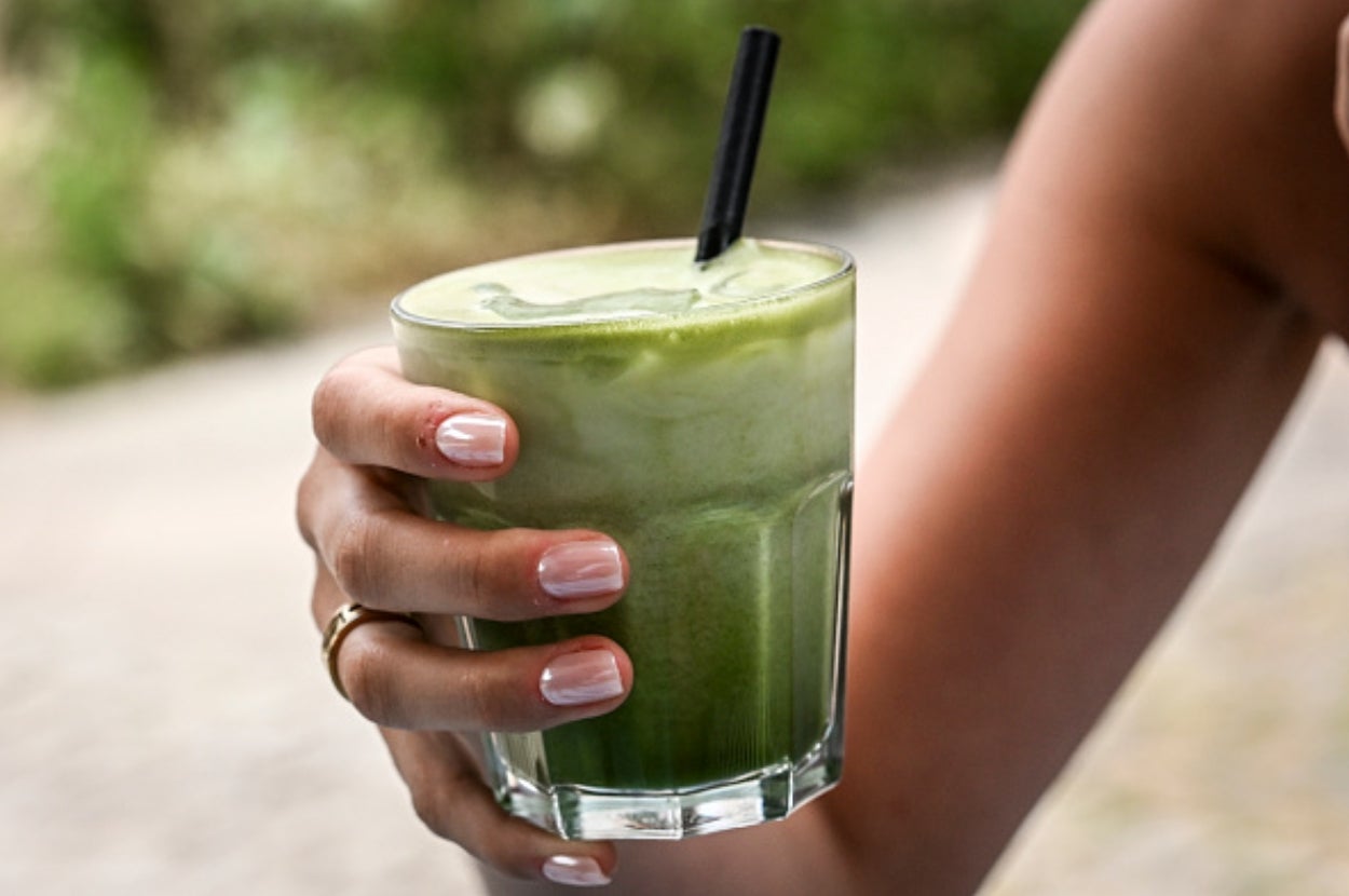 Person holding a glass of green smoothie with a black straw, outdoors