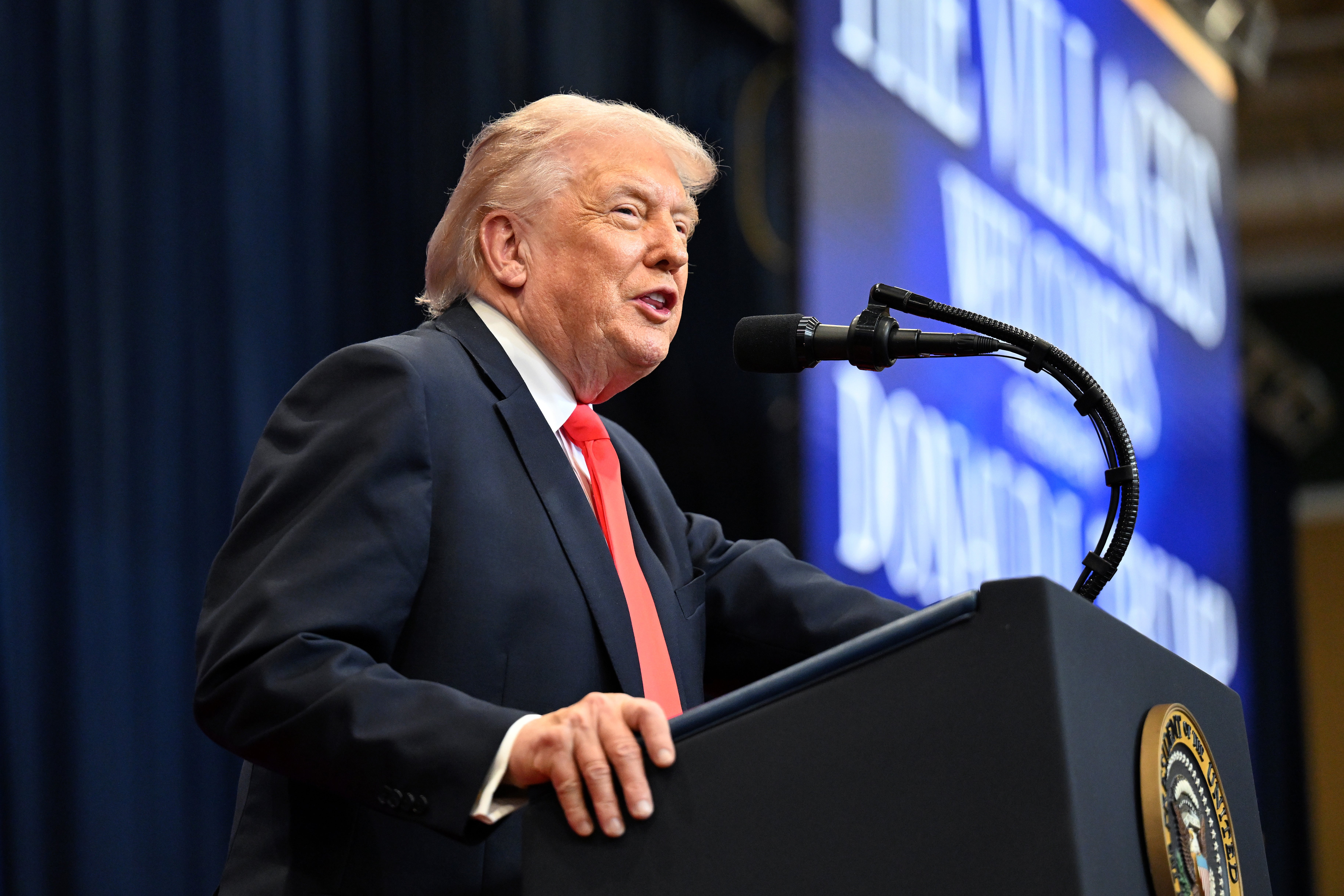 Man astatine  podium speaking astatine  an event, wearing a acheronian  suit   and reddish  tie, with a blurred inheritance  displaying partial text