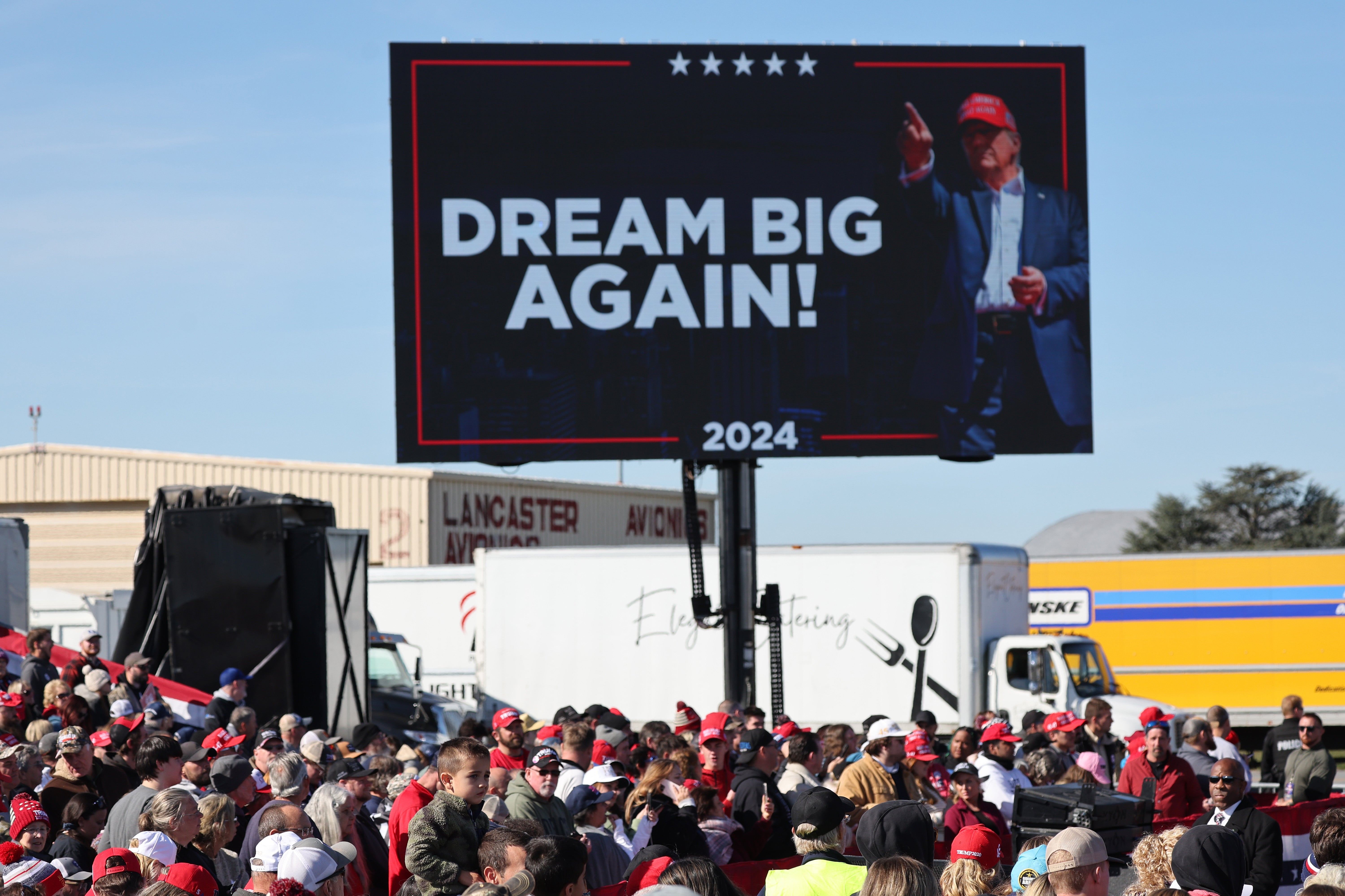 Crowd gathers outdoors nether  a ample  billboard showing a antheral   successful  a suit   and chapeau  with the words "Dream Big Again! 2024."