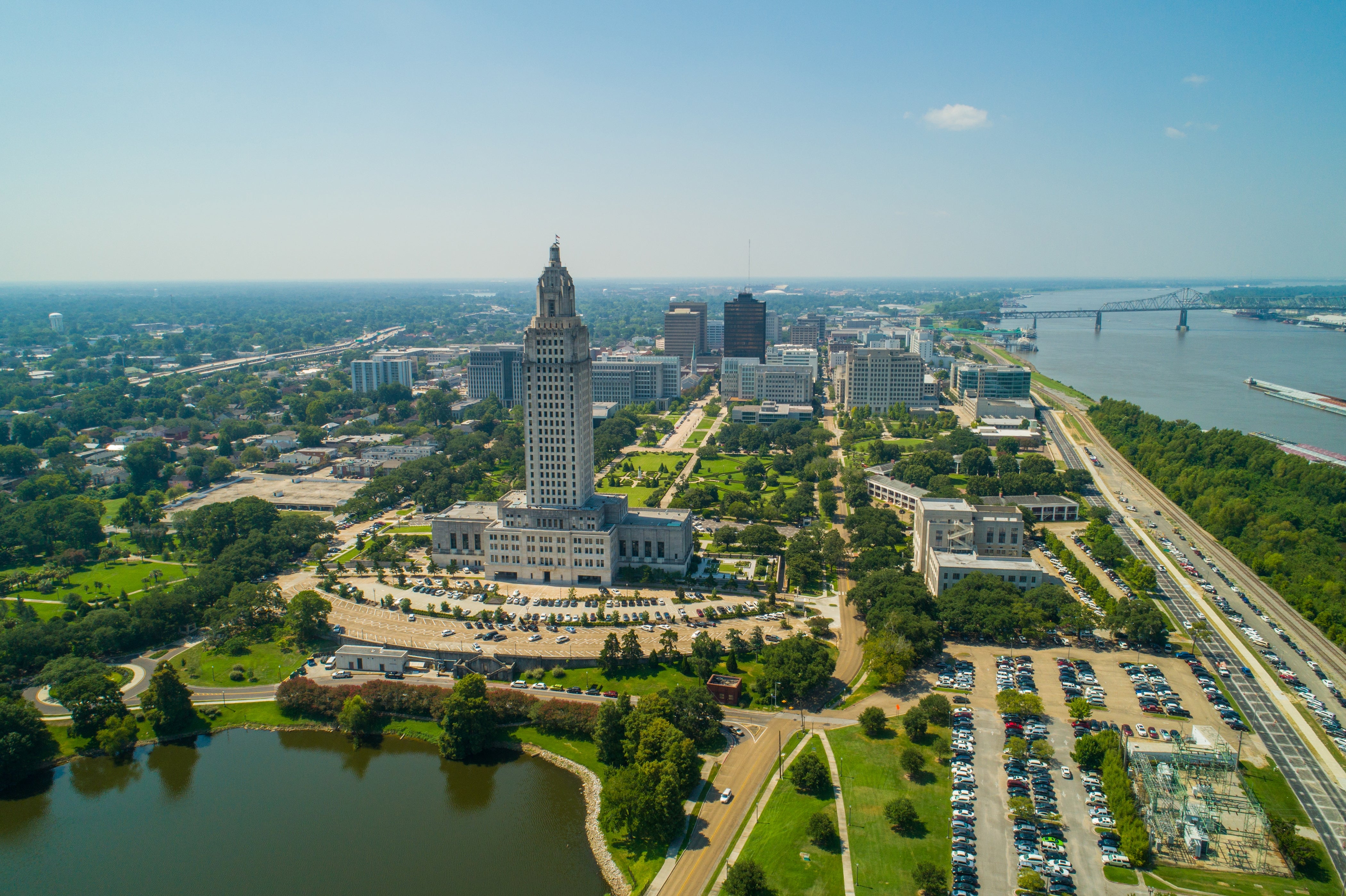 Aerial presumption    of Baton Rouge, featuring the Louisiana State Capitol building, surrounded by greenery and the Mississippi River connected  the right