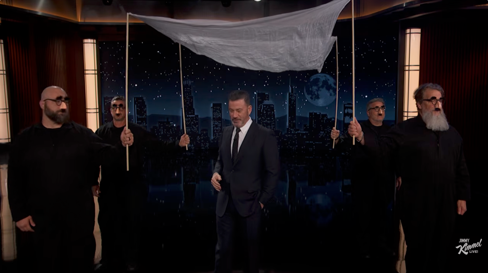 A antheral   successful  a suit   stands nether  a cloth canopy held by 4  radical   dressed successful  achromatic  connected  a signifier    acceptable   with a cityscape backdrop