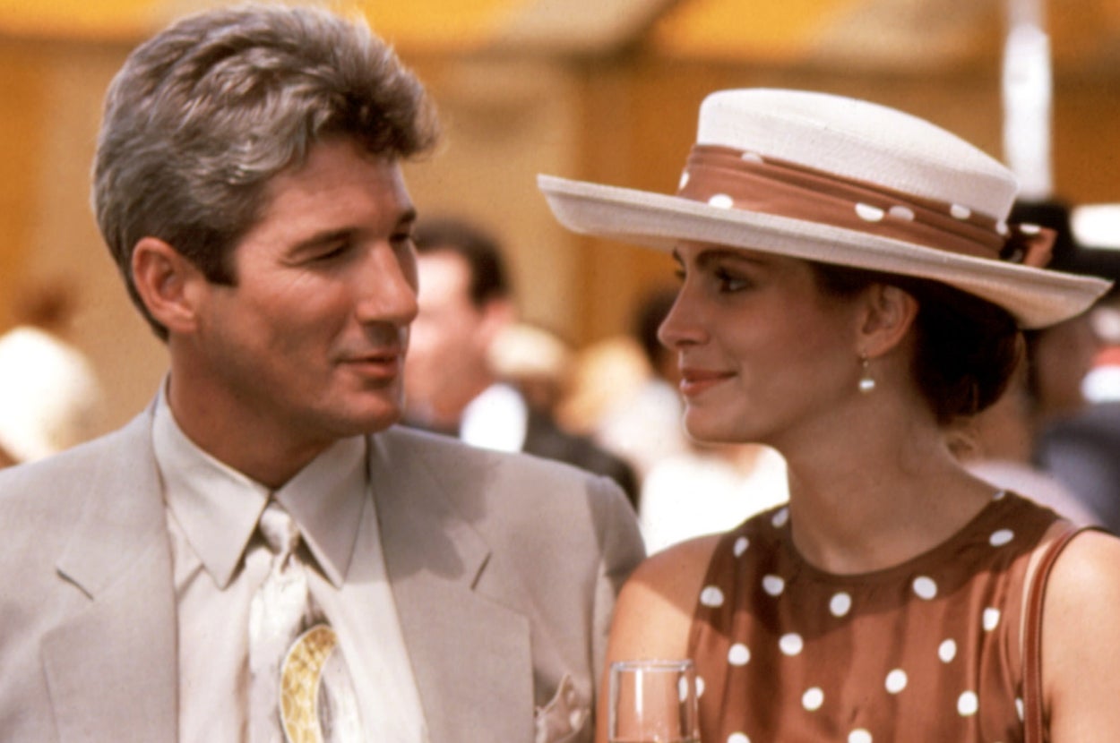 A man in a suit and a woman in a polka dot dress and hat converse, sharing a gentle moment at an outdoor social event