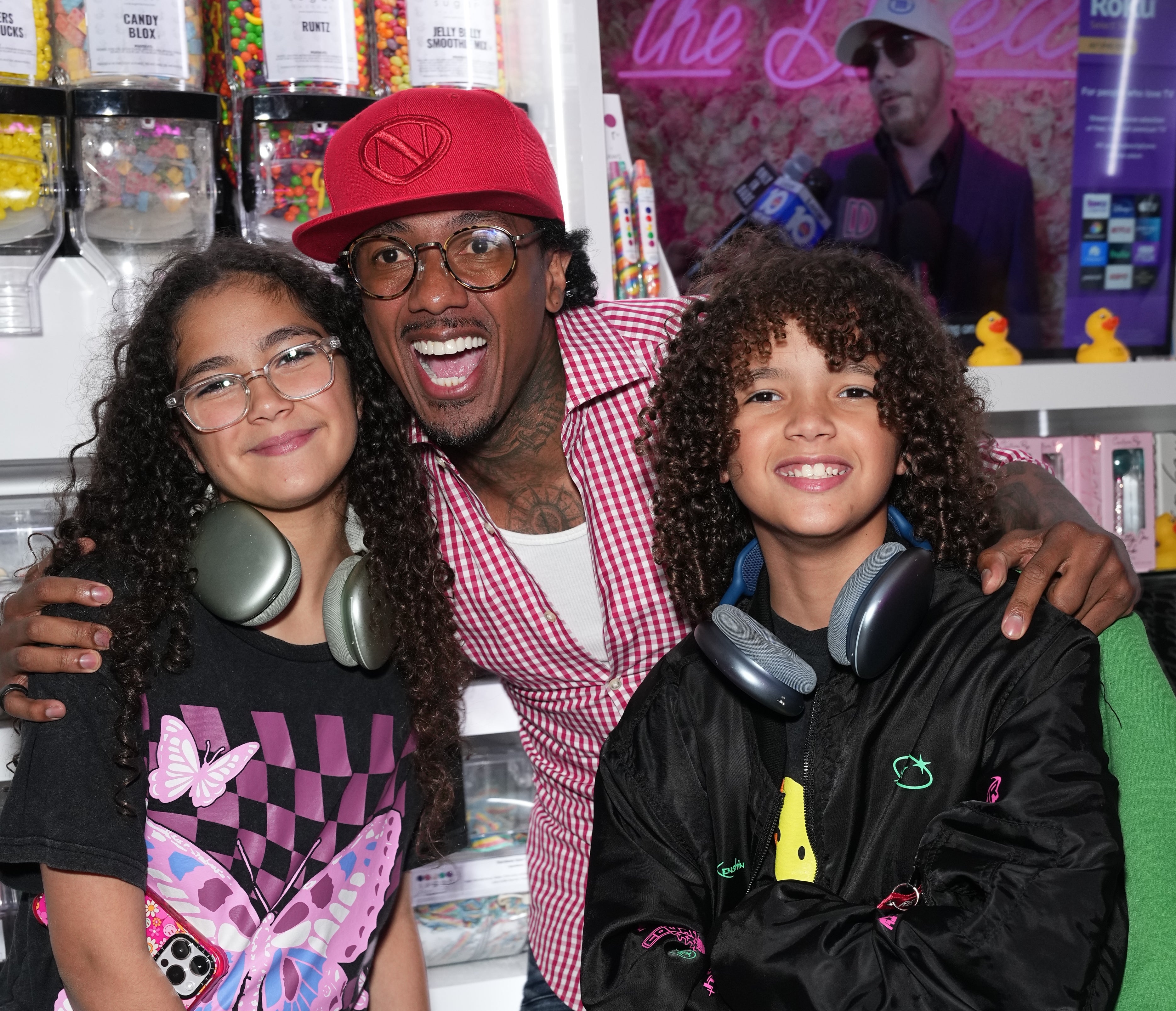 A idiosyncratic   with agelong  curly hairsbreadth  stands smiling betwixt  2  children wearing headphones, successful  a candy store   setting