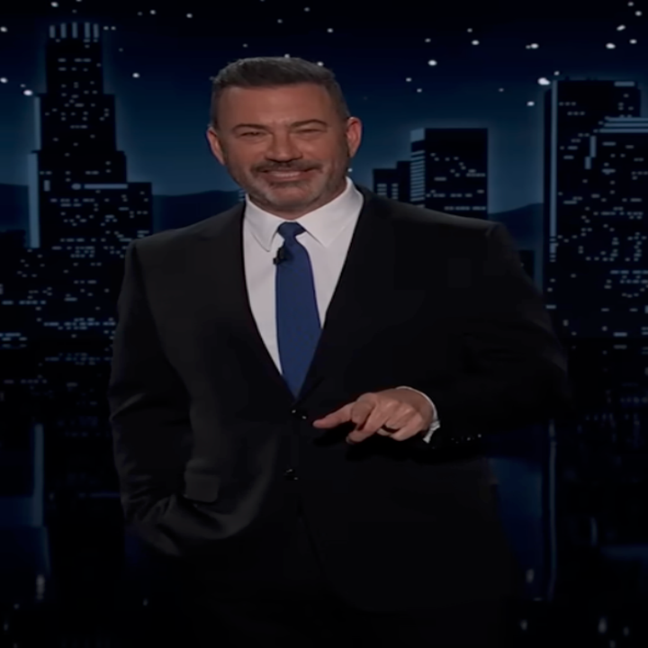 A man in a suit and tie stands against a city skyline backdrop on a nighttime talk show set