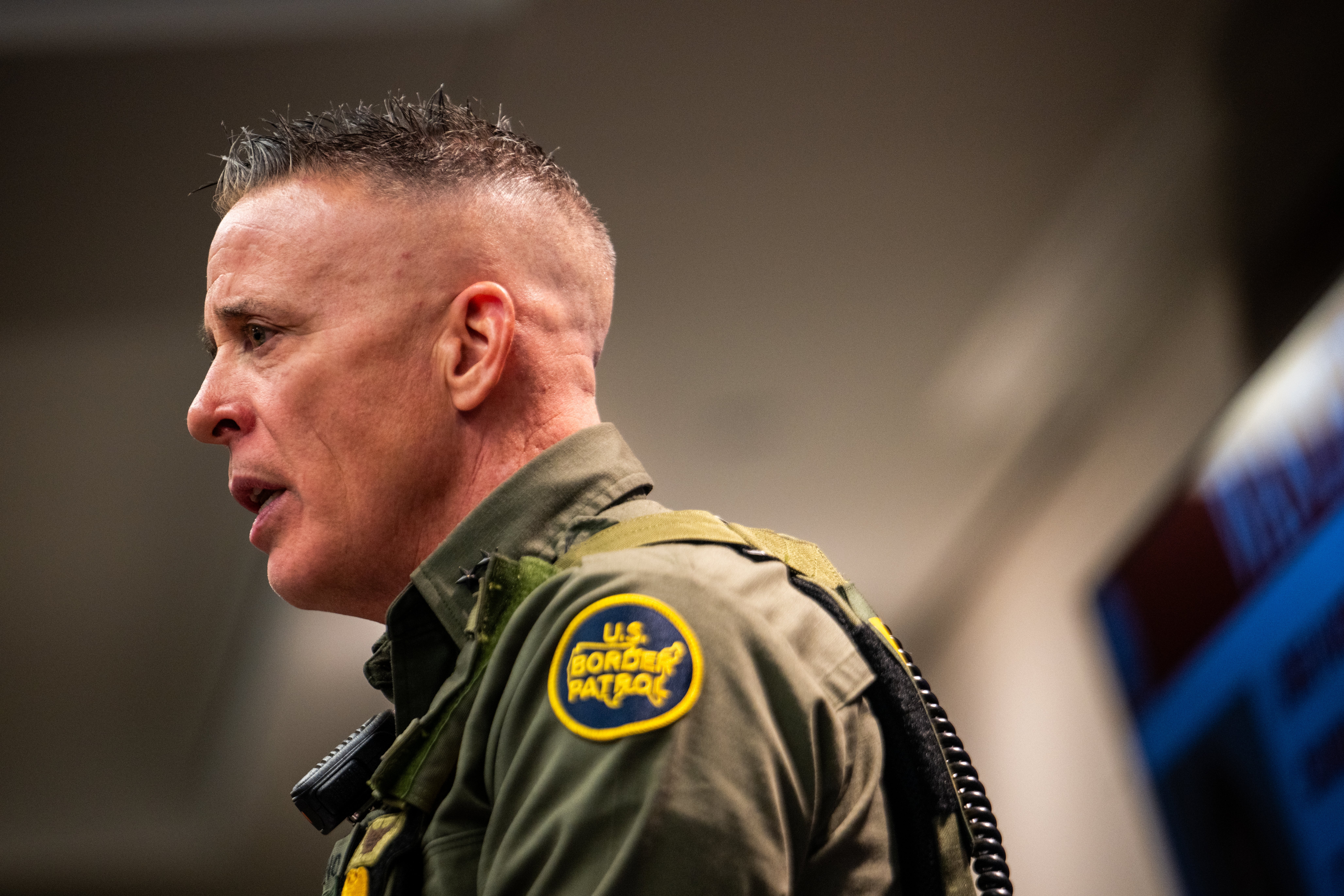 En amerikansk grensepatruljeoffiser snakker på et innendørsarrangement, iført uniform med en synlig byrålapp på skulderen