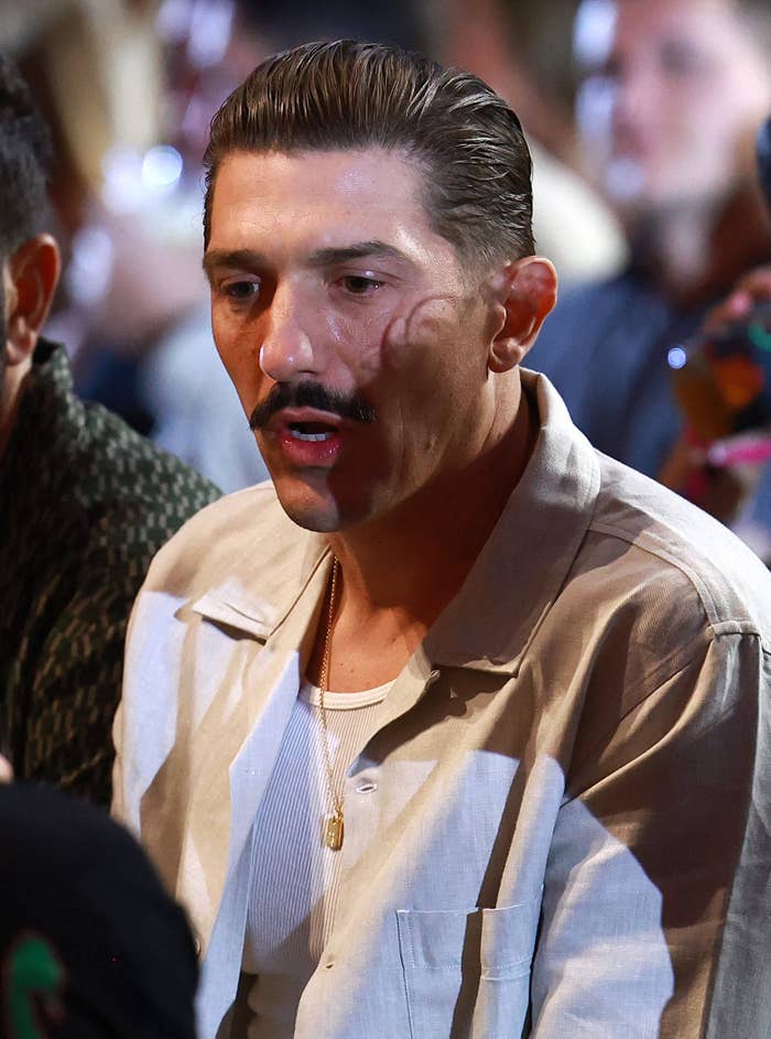 Man with slicked-back hair and mustache wearing a beige jacket over a white shirt at an event