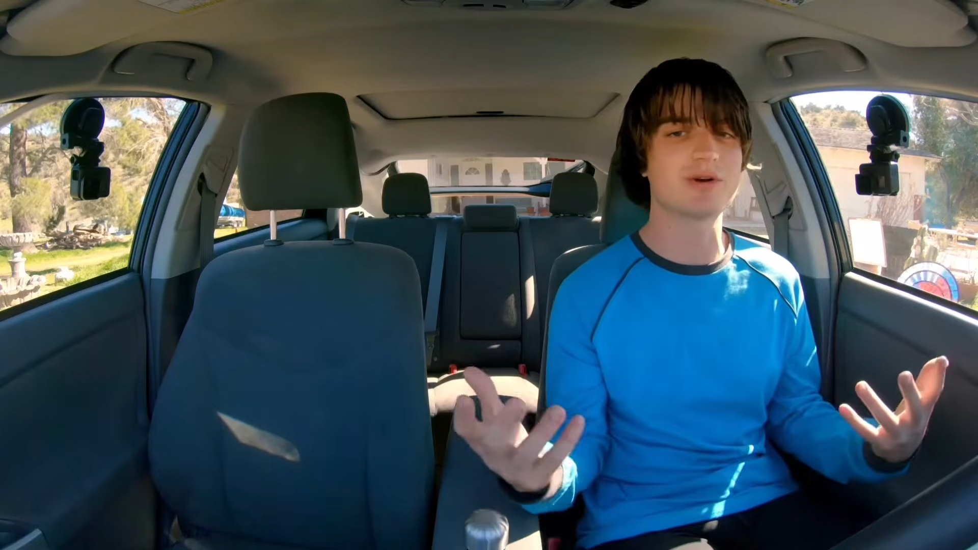 A person gestures while sitting in a car's driver seat, appearing to be speaking or singing