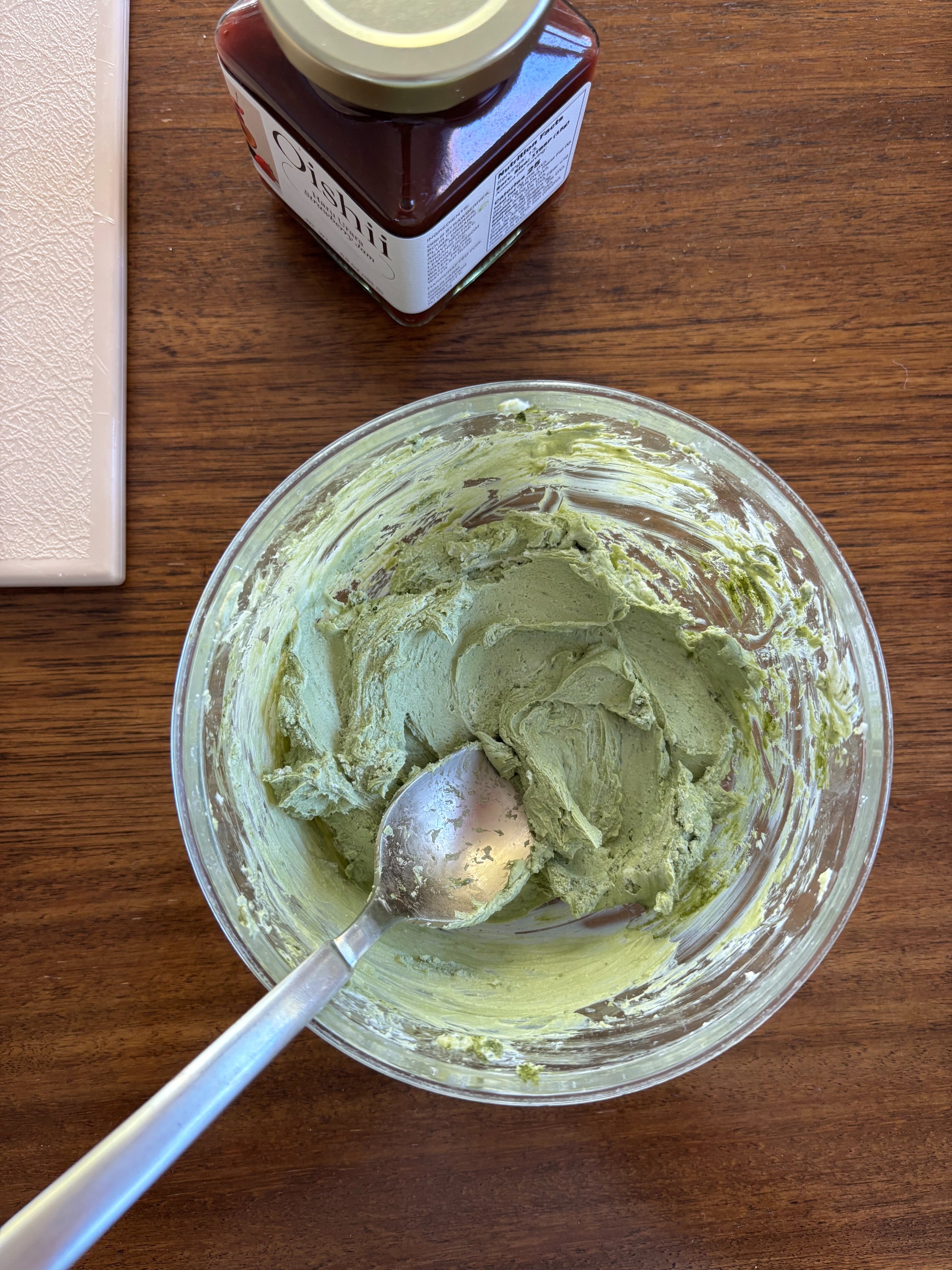 Bowl with matcha spread and a spoon on a wooden table, jar of berry jam nearby