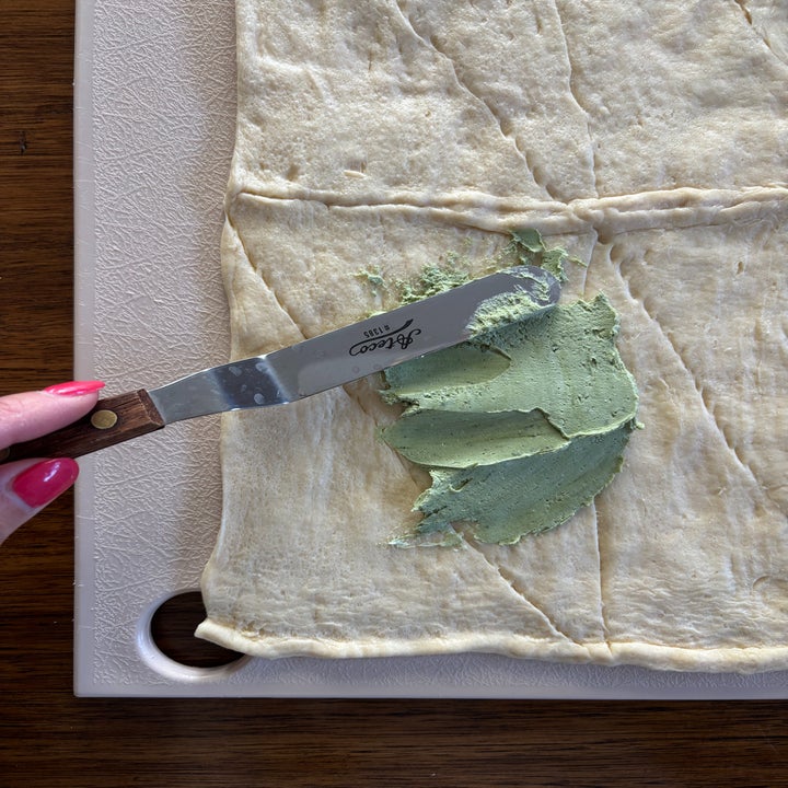 Hand holding offset spatula, spreading green filling onto piece of dough