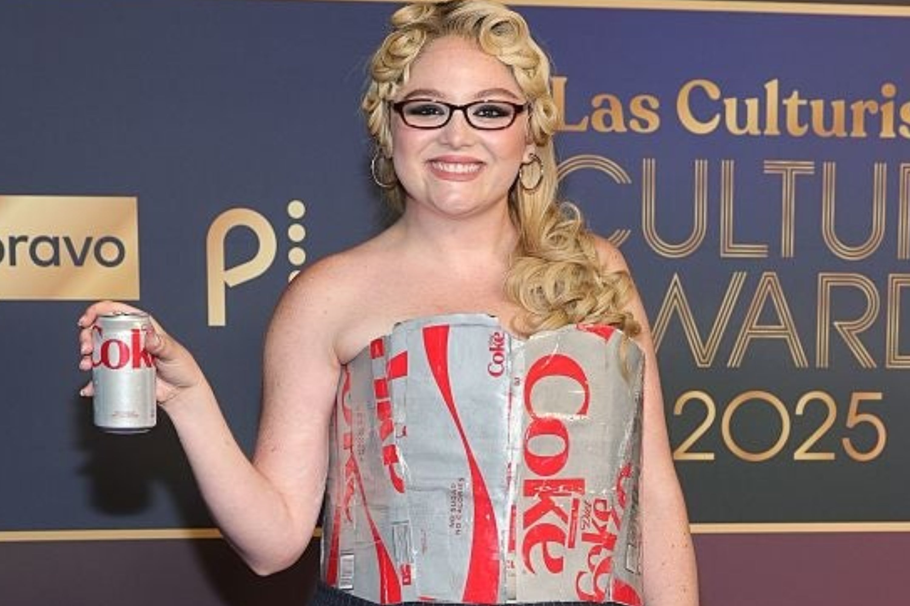 Person in a strapless dress made from soda can boxes, holding a diet soda can, at an awards event backdrop