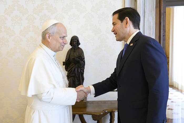 A man in a white religious robe shakes hands with another man in a suit in an ornate room with a statue in the background