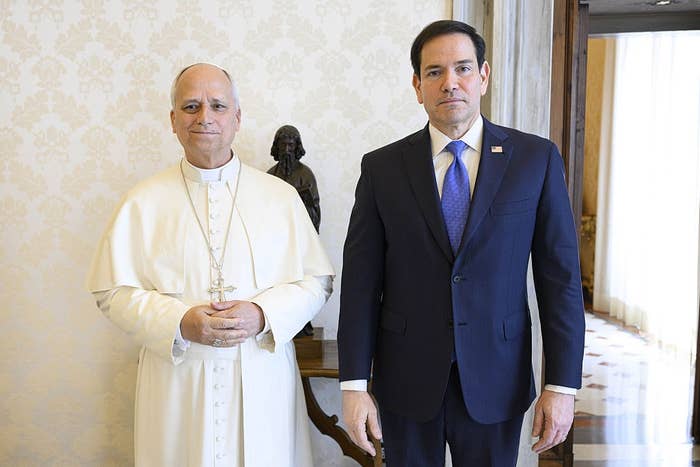 Two individuals in formal attire stand side by side, one in religious robes and the other in a suit with a tie