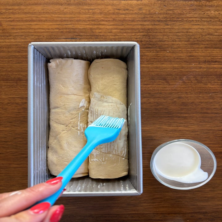 Two logs of unbaked dough in a loaf pan are being brushed with cream