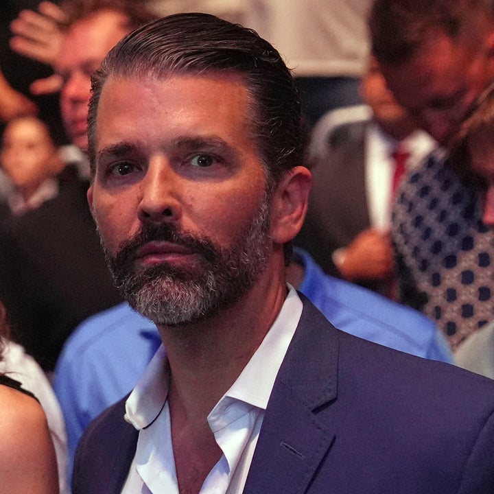 A man in a suit and a woman in a sleeveless dress stand in a crowded event, looking towards the camera