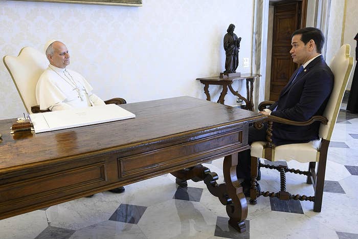 Two individuals sit across from each other at a wooden desk in a formal setting, with a statue in the background