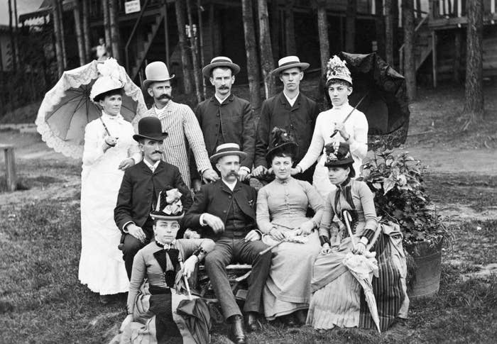 Victorian-era radical  representation    with men successful  suits and bowler hats, women successful  elaborate dresses and hats, posing outdoors with parasols