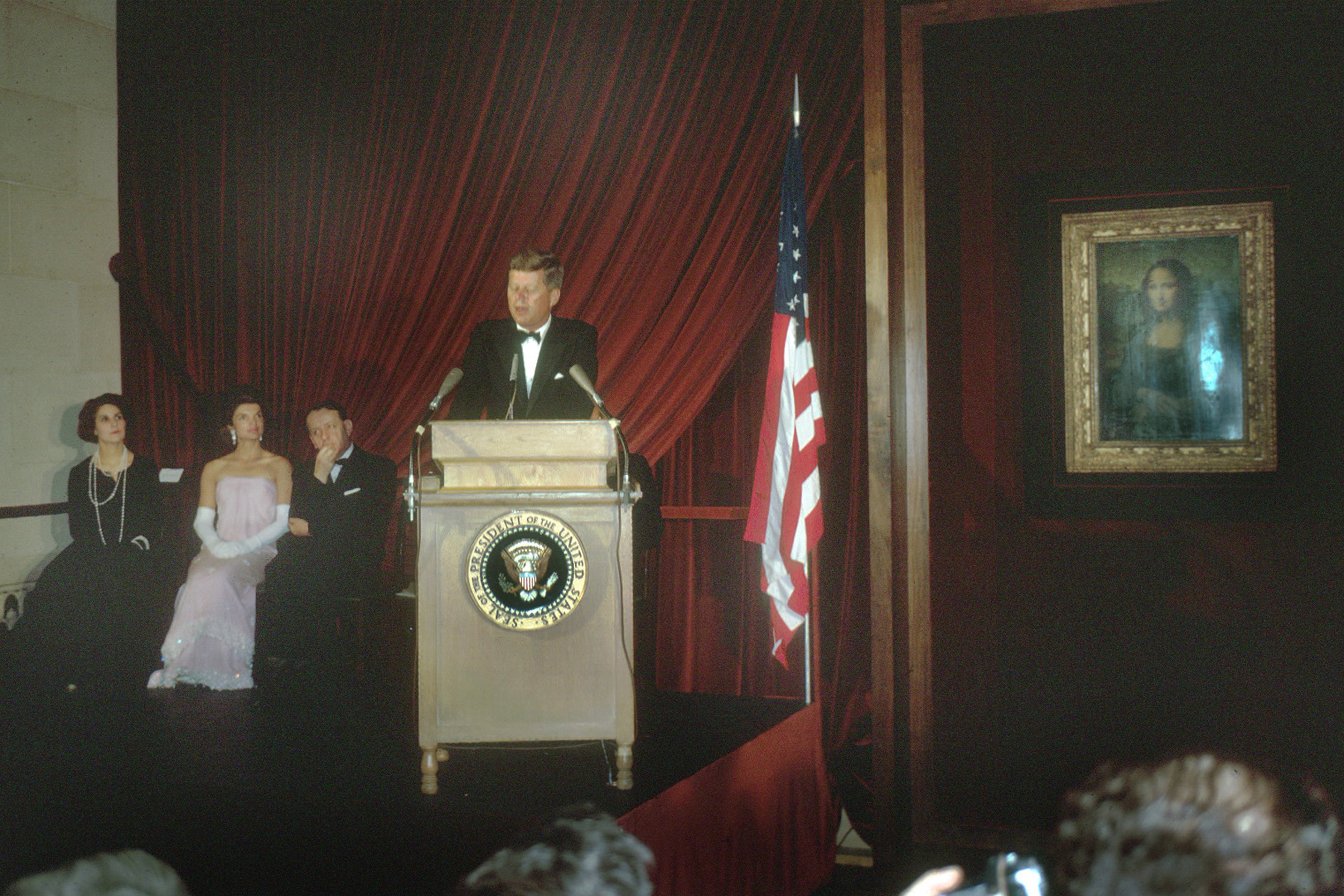 A antheral   successful  a tuxedo speaks astatine  a podium with the U.S. statesmanlike  seal, flanked by seated individuals and a U.S. flag. A representation    hangs nearby
