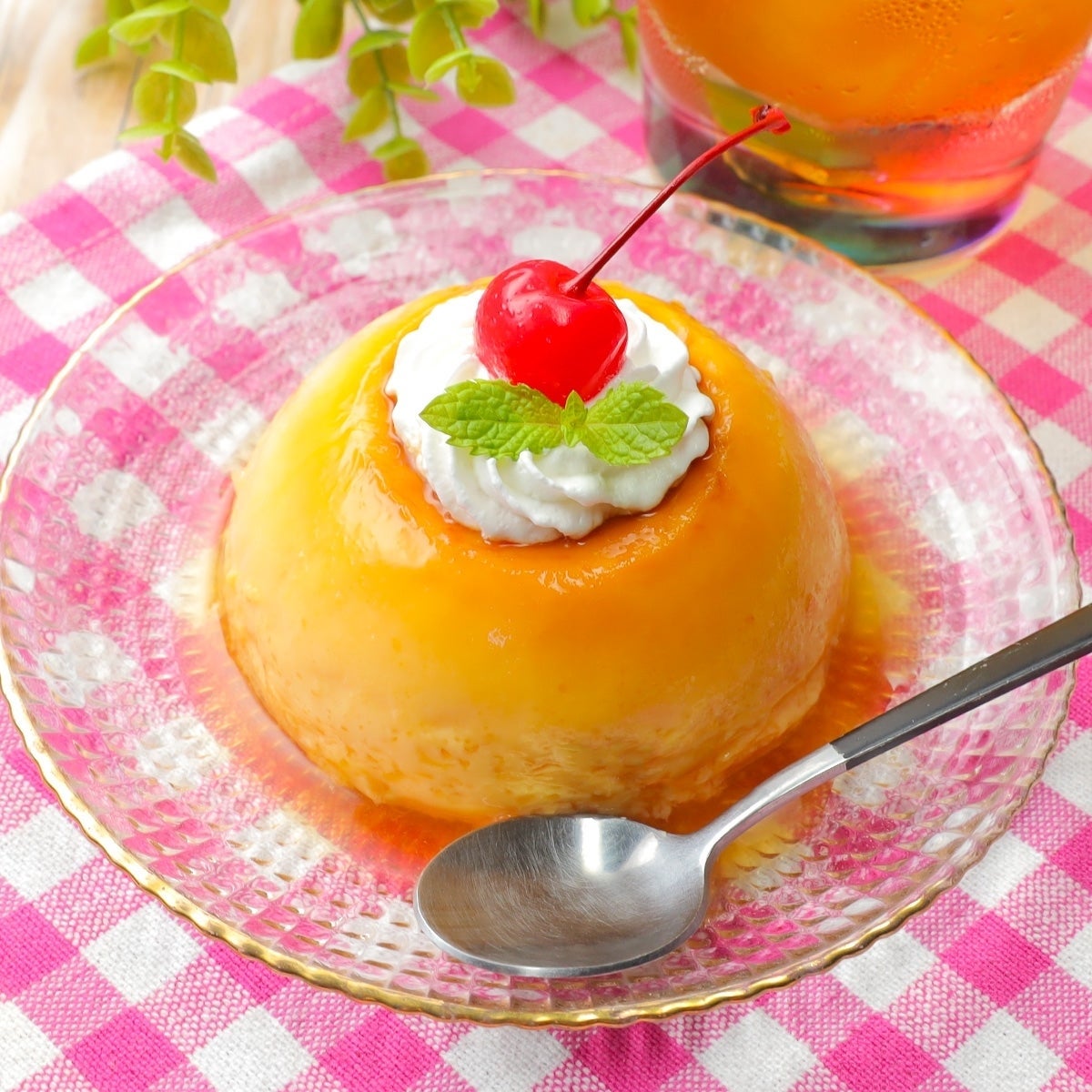 カスタードプリンにホイップクリームとチェリーがトッピングされ、ミントの葉が添えられたデザート。隣にスプーン付き。