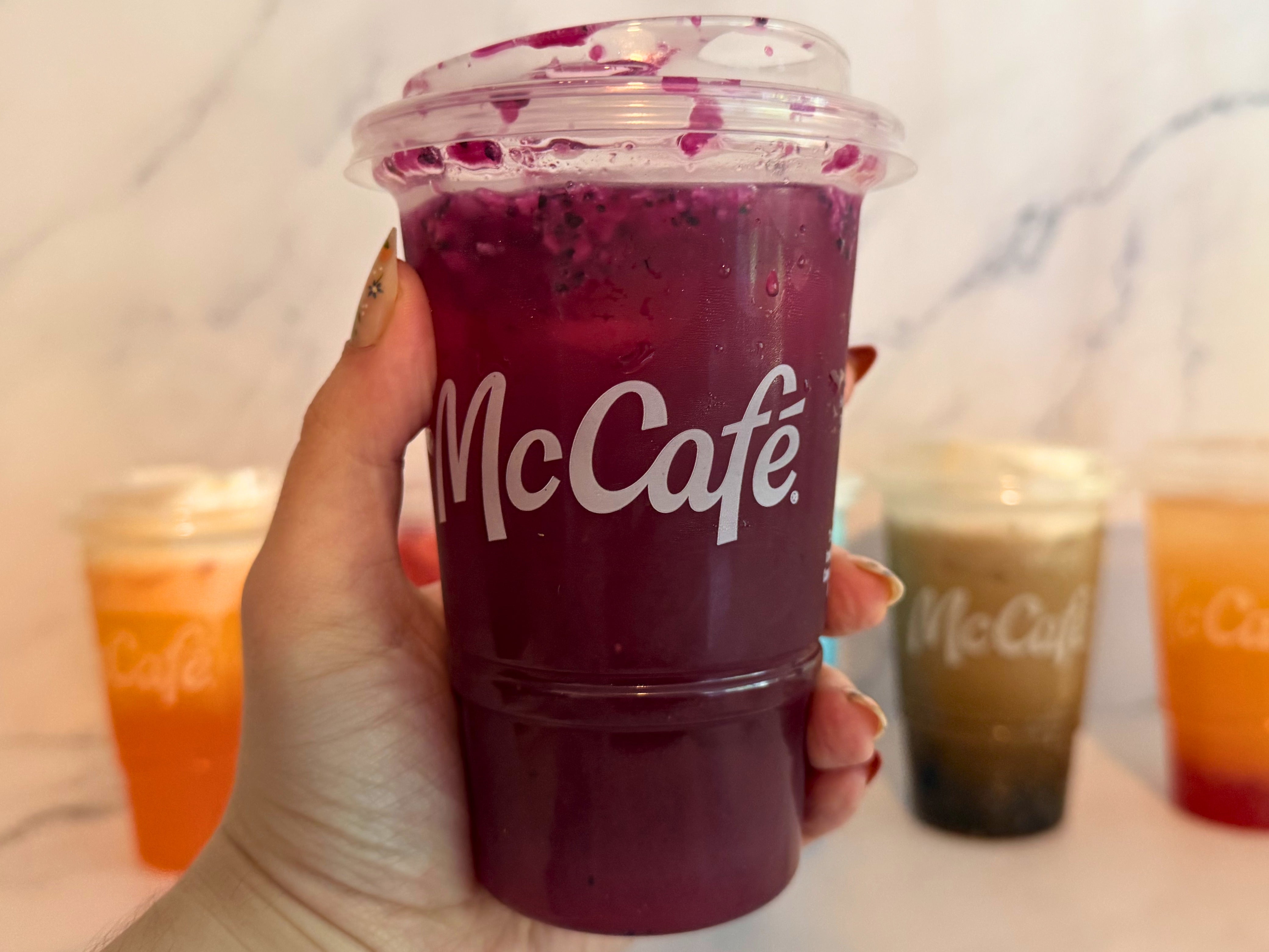 A hand holds a McCafé Blackberry Passion Fruit refresher with other colorful drinks blurred in the background