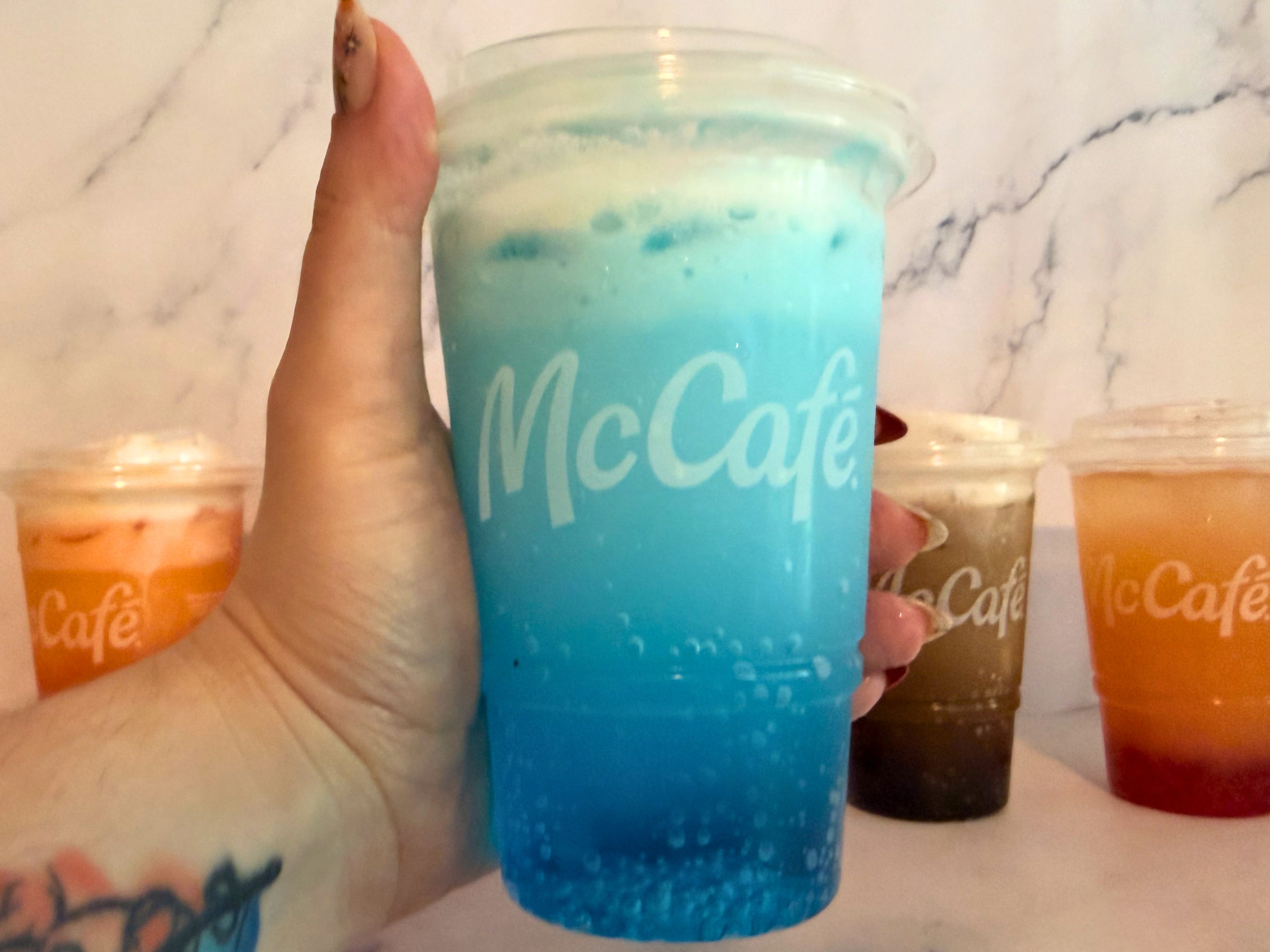 Hand holding a Sprite Berry Blast in a McCafé cup, with other colorful drinks in McCafé cups in the background