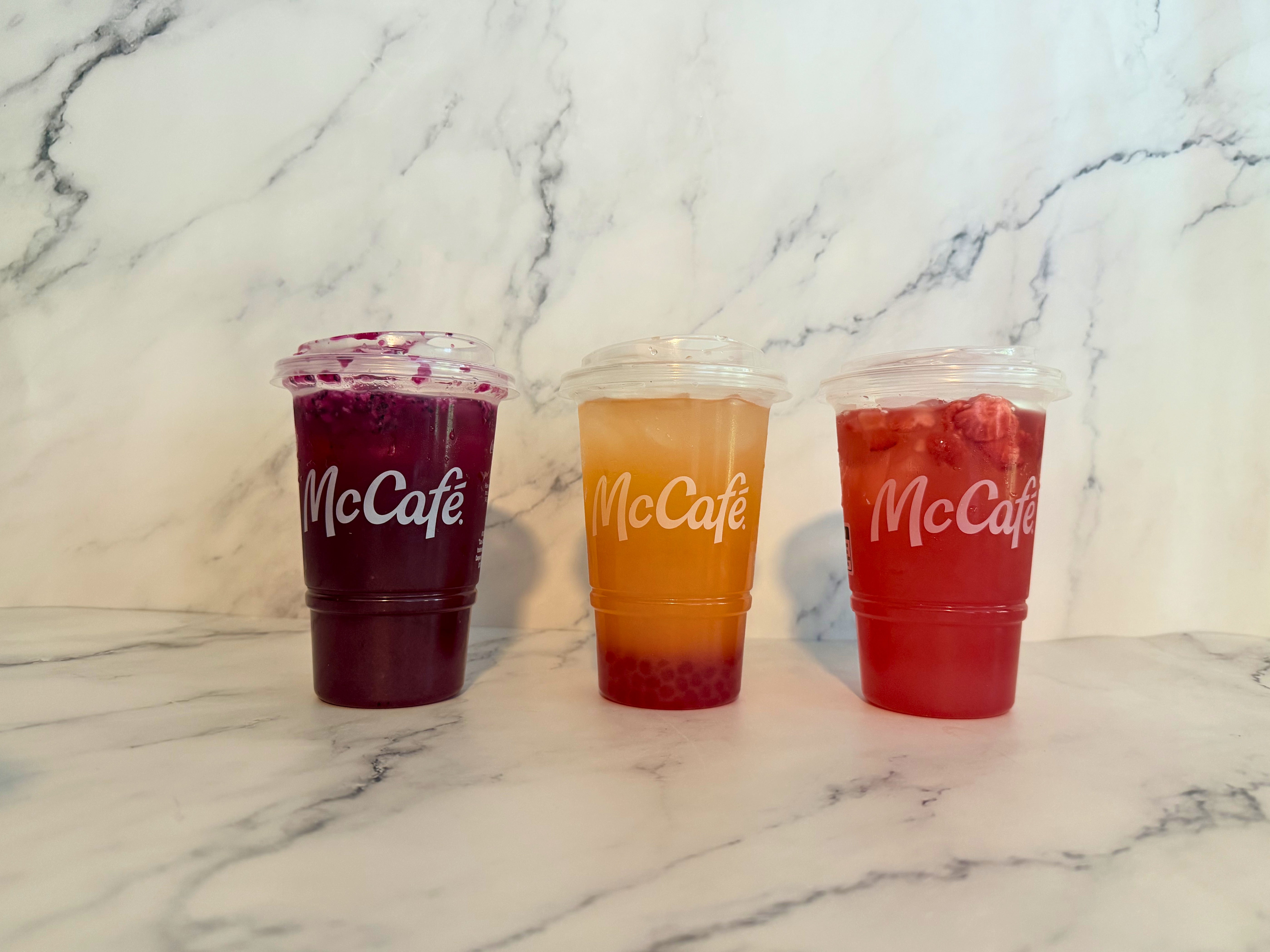 Three McCafé refreshers on a marble surface: purple with berries, orange with bubbles, and red with fruit pieces