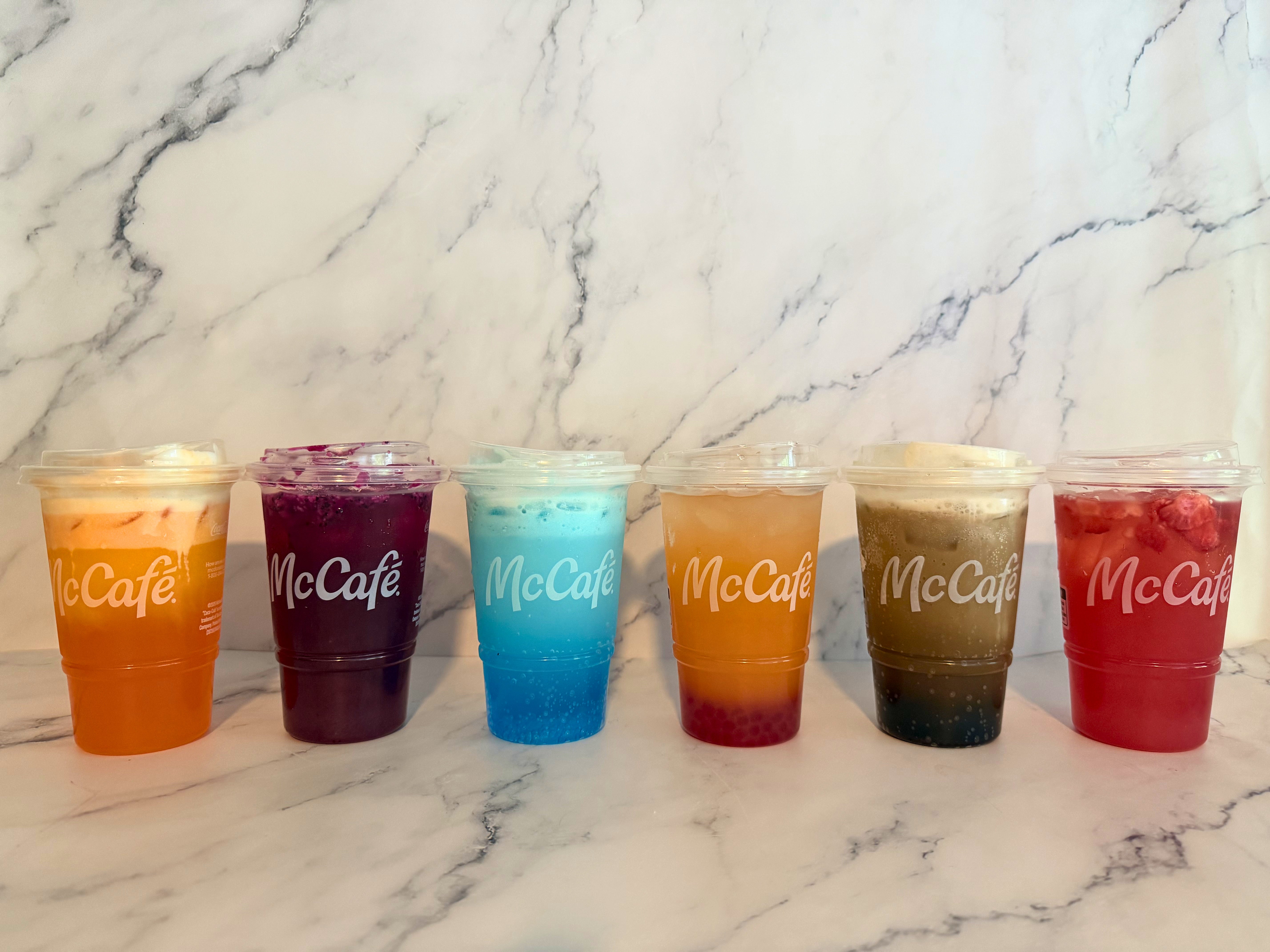Six McCafé dirty sodas and refreshers in transparent cups with lids, showcasing a variety of beverages on a marble-like surface