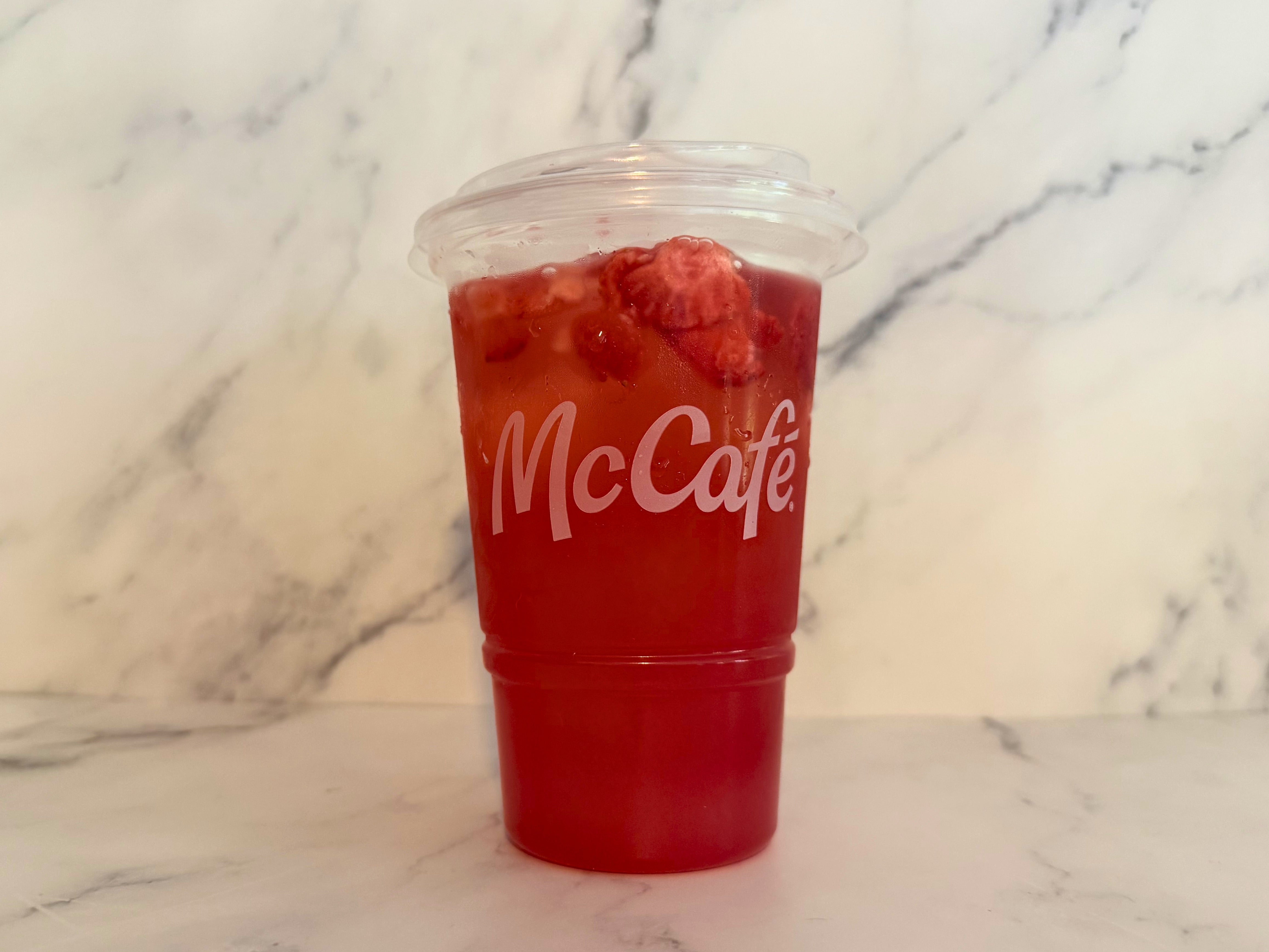 A plastic cup with a McCafé logo is filled with a Strawberry Watermelon Refresher, topped with strawberry slices, set against a marble background
