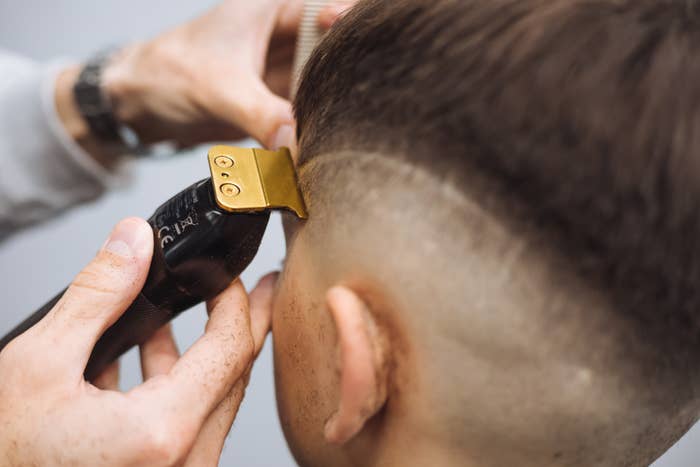 Person getting a haircut with electric clippers, showing a close-up of the barber's hands and a design being shaved into the hair
