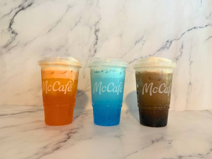 Three McCafé iced drinks of different flavors are on a marble surface, each in a plastic cup with a dome lid
