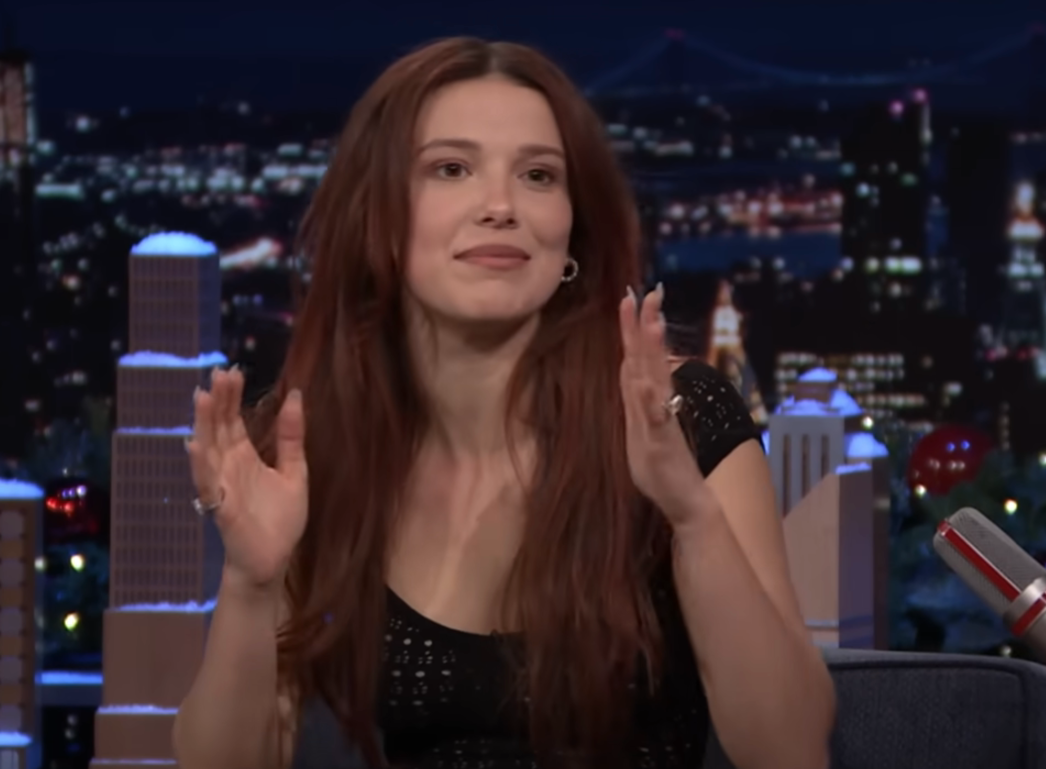 Millie Bobby Brown gesturing animatedly during a nighttime talk show with Jimmy Fallon