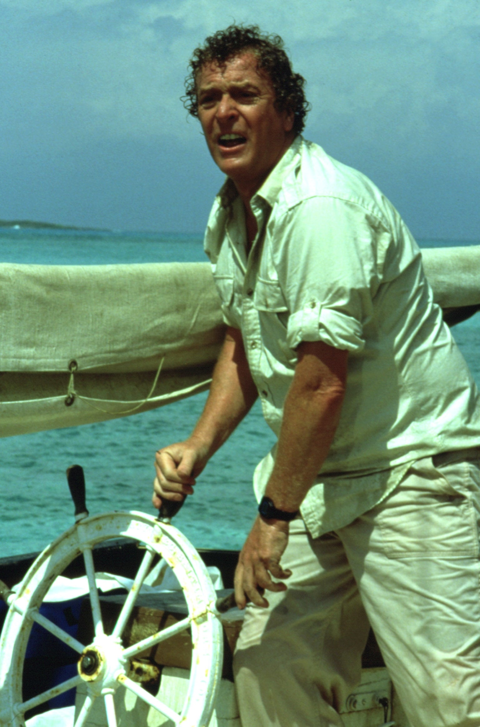 Person steering a boat's wheel, wearing a casual button-up garment  and airy  pants, against a backdrop of the sea