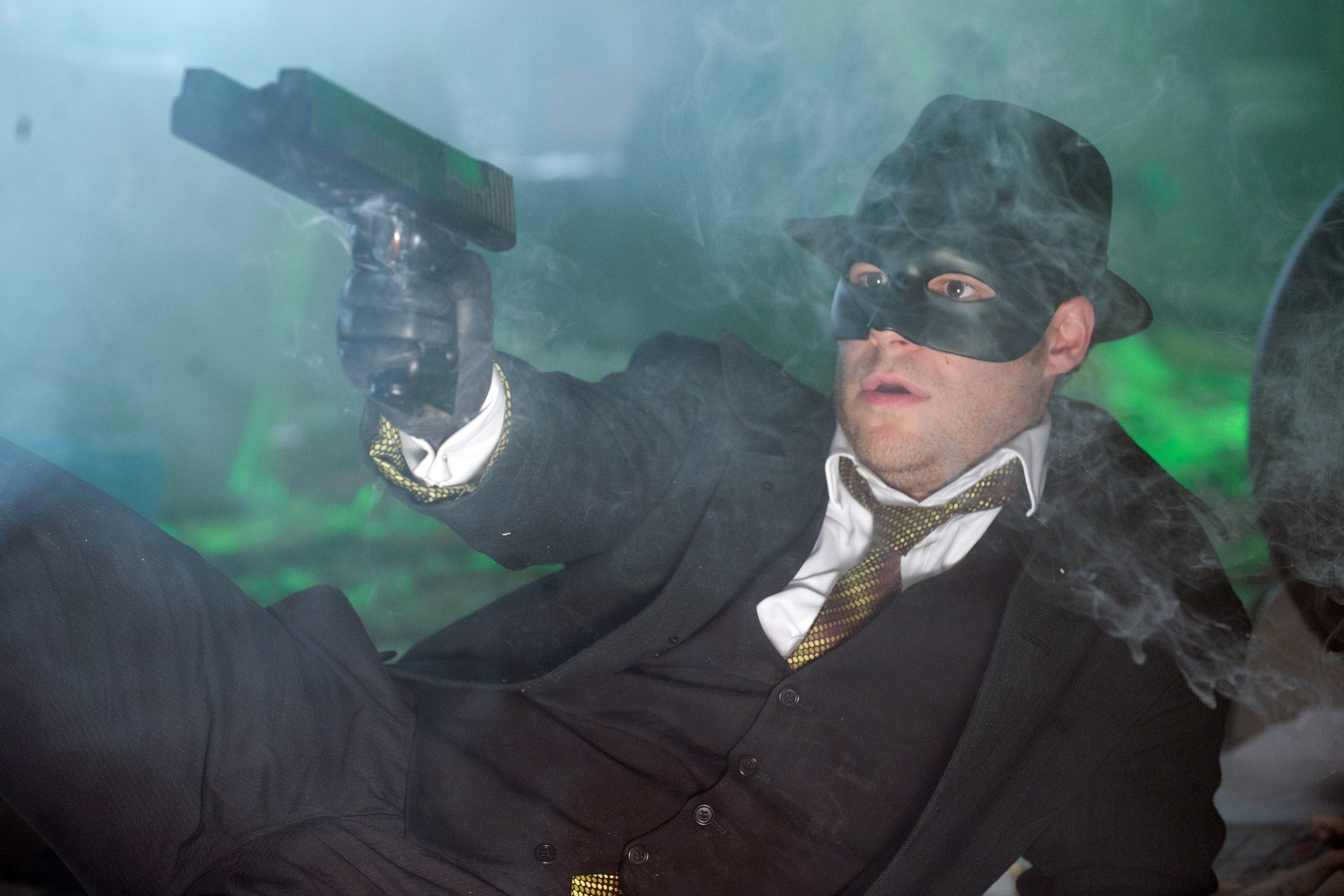 Person successful  a suit, tie, and hat, wearing a achromatic  mask, holds a weapon  amid smoke, creating a melodramatic  scene