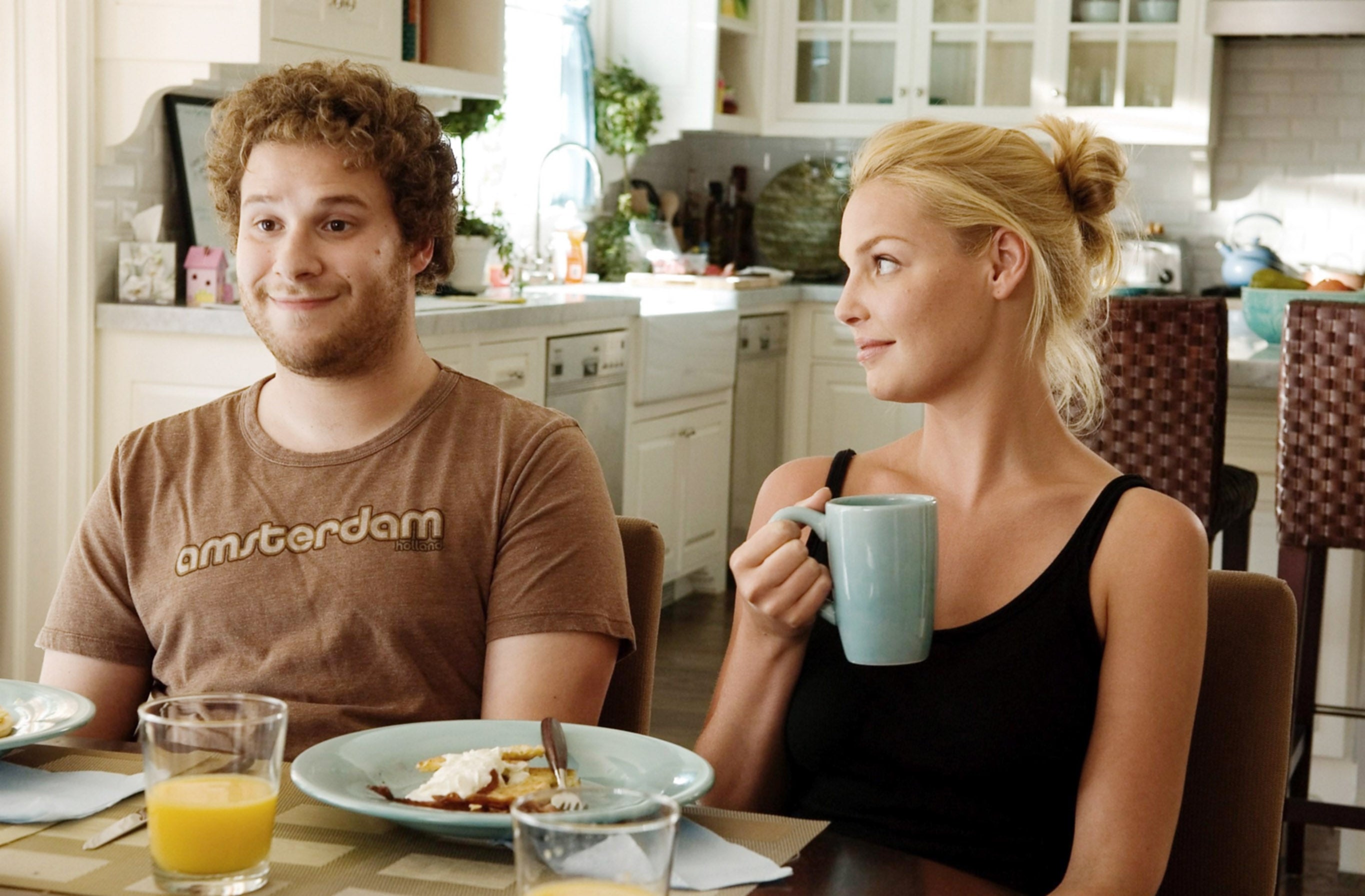 Two radical   sitting astatine  a room  table; 1  holds a mug, looking astatine  the different   who appears amused. Plates and drinks are connected  the table