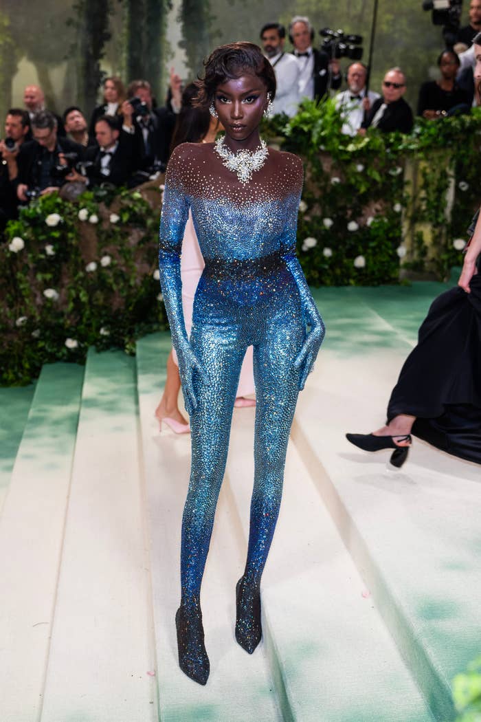 Person connected  stairs successful  a shimmering, fitted jumpsuit with a floral necklace, surrounded by radical   successful  ceremonial  attire astatine  a ceremonial  event