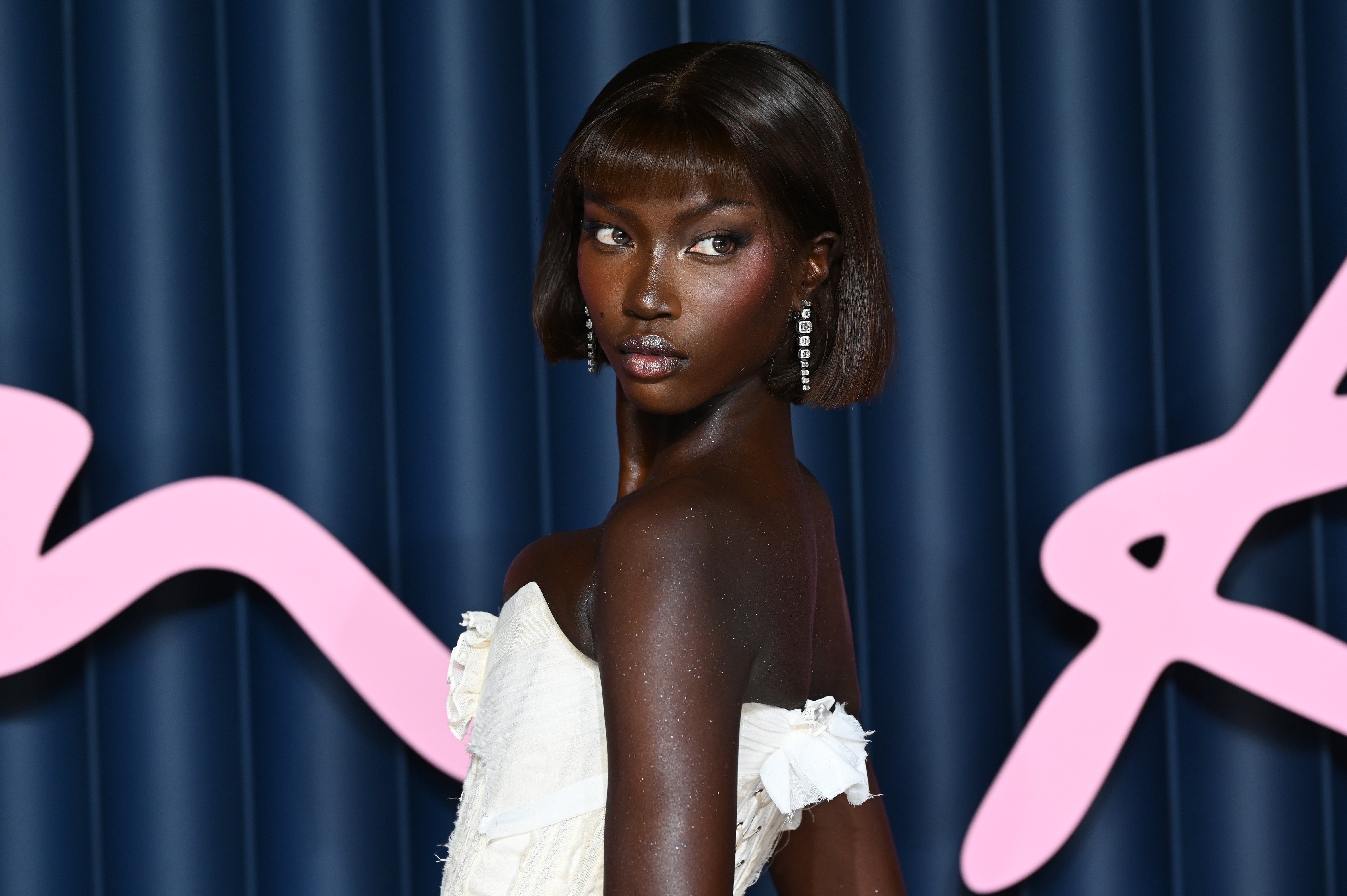Anok Yai successful elegant, strapless gown with earrings, posing against a bluish backdrop with abstract pinkish lines