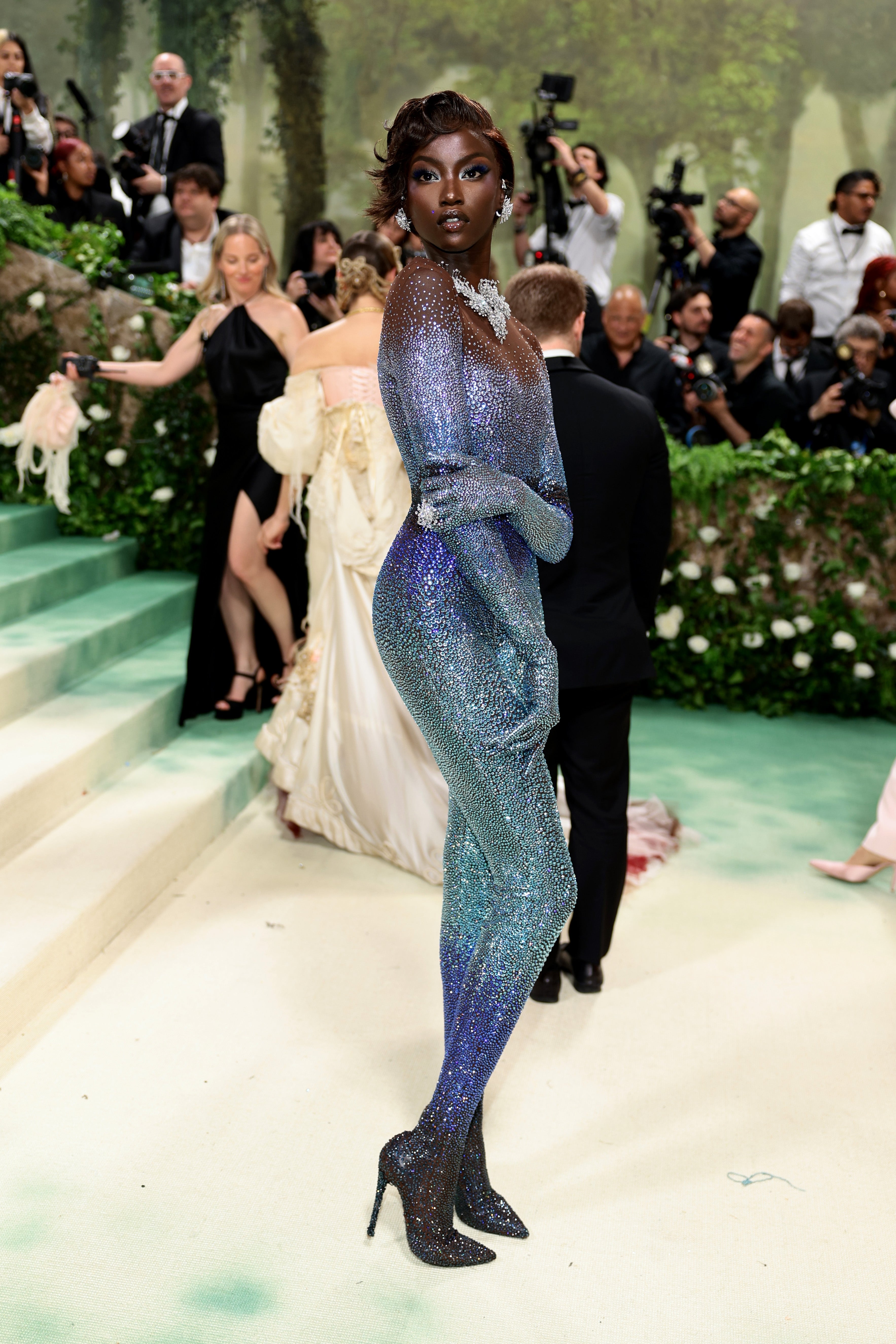 Anok Yai l successful a sparkling full-body outfit with a connection necklace poses elegantly connected a high-profile lawsuit carpet, surrounded by photographers and guests