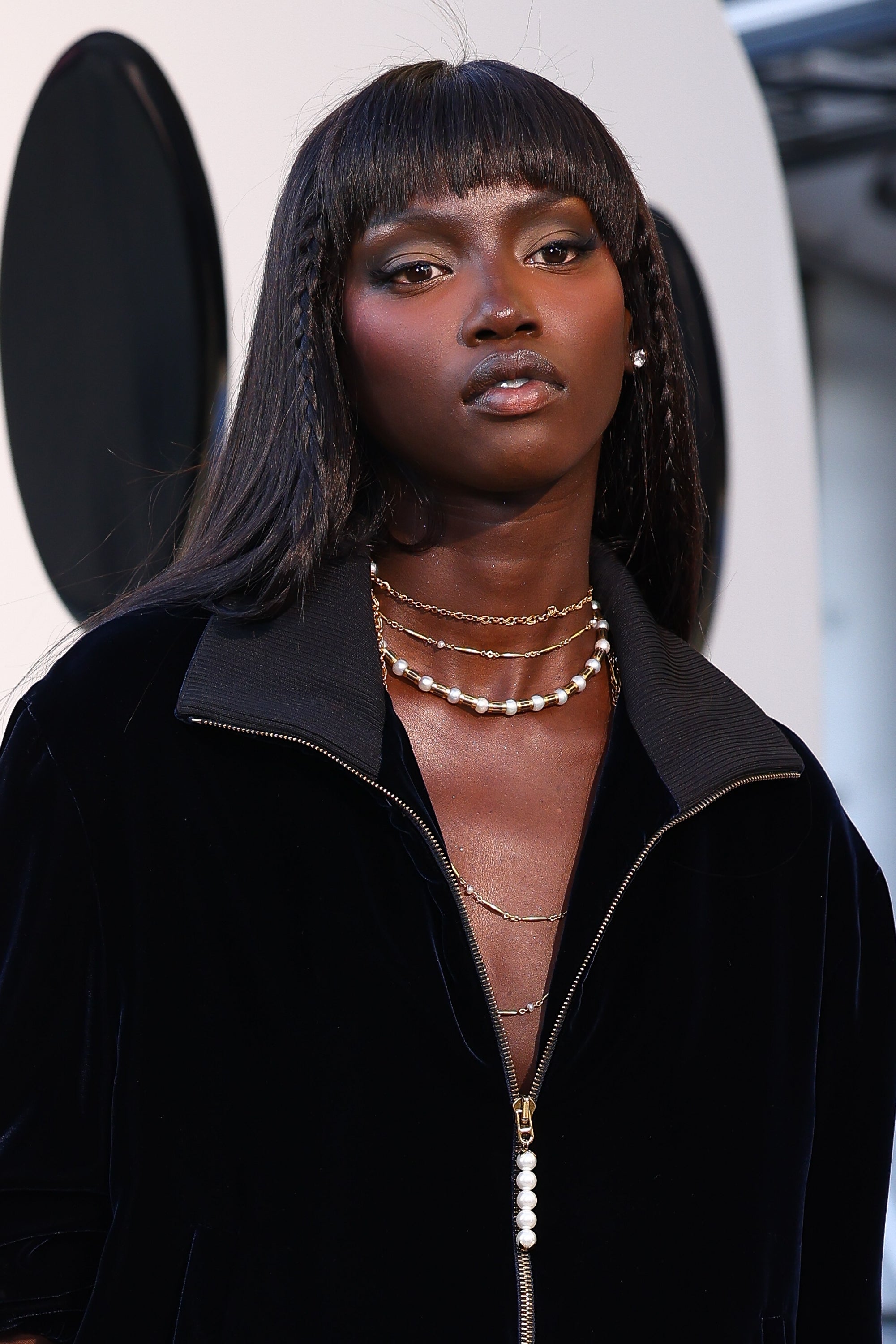 Anok Yai wearing a sleek overgarment with layered necklaces stands against a neutral backdrop
