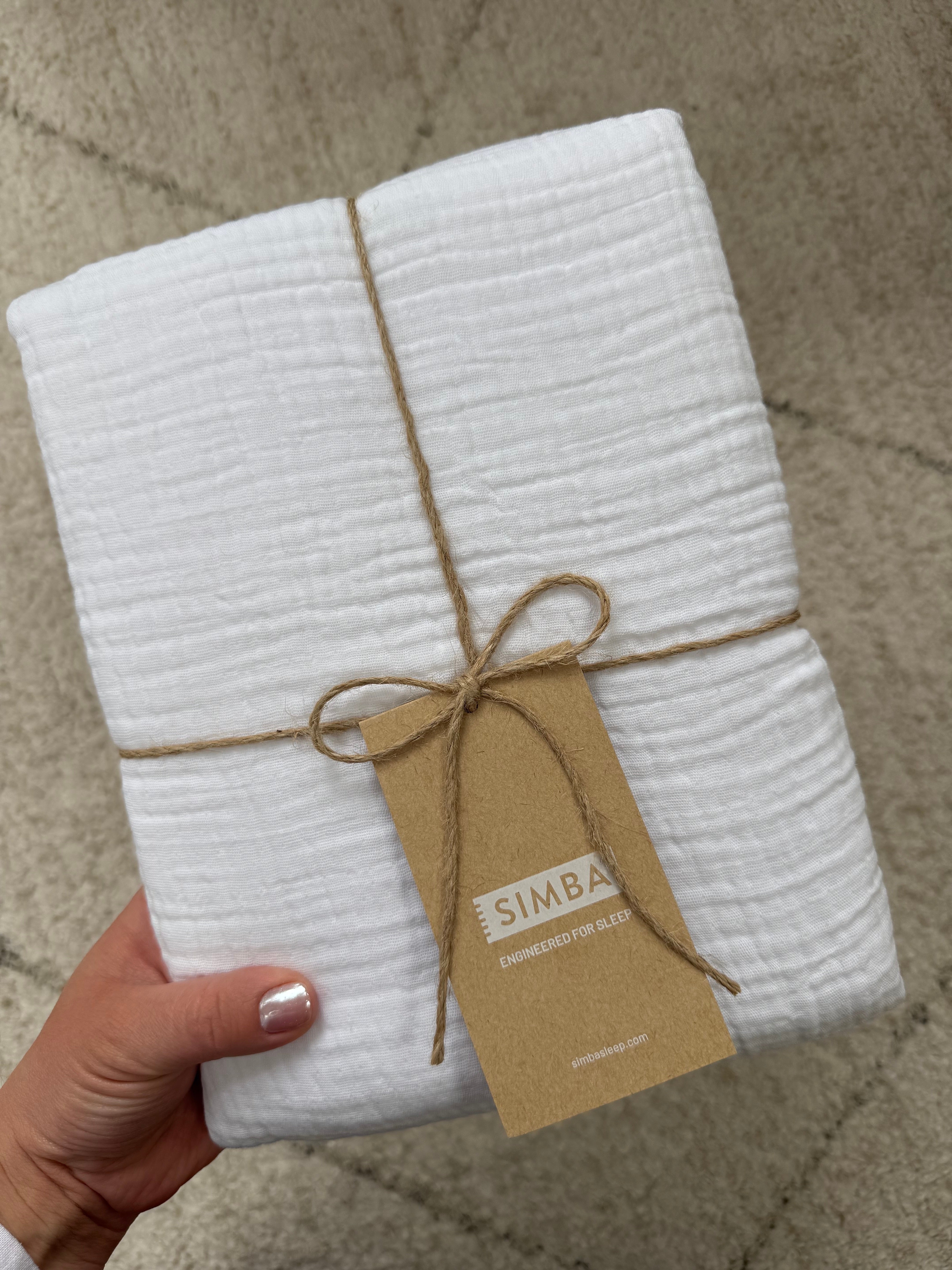 Orang yang memegang bungkusan kain bertekstur putih yang terbungkus rapi, diikat dengan pita benang sederhana, dengan label berlabel “Simba.”