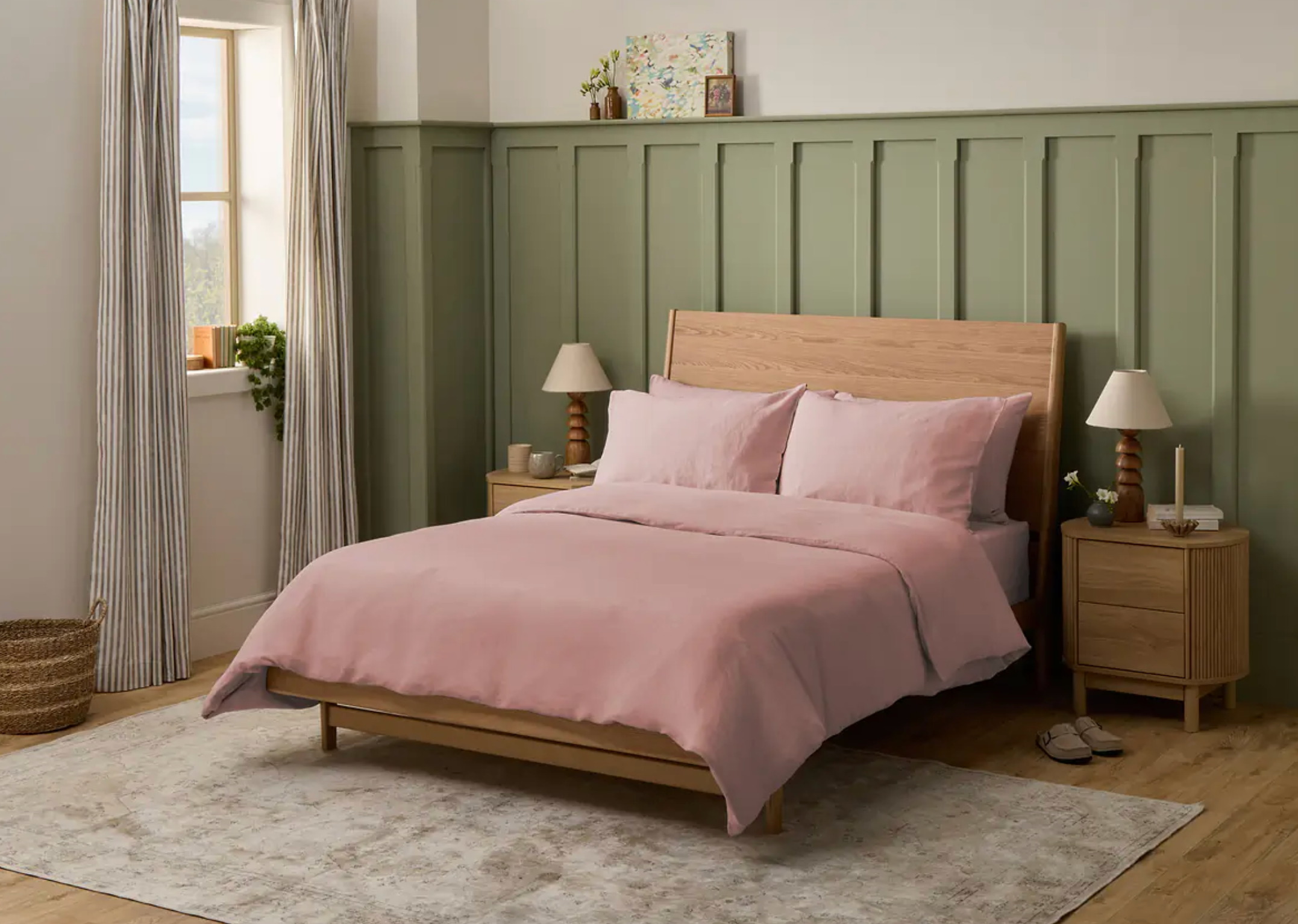 Kamar tidur dengan rangka tempat tidur kayu, tempat tidur merah muda, meja samping dengan lampu, dan dekorasi. Kamar ini memiliki jendela dengan tirai dan dinding beraksen berpanel hijau