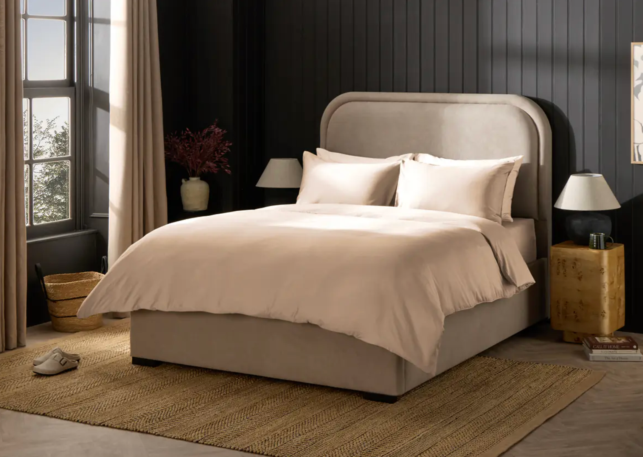 Penataan kamar tidur yang elegan dengan tempat tidur mewah, dua bantal, tempat tidur netral, lampu samping tempat tidur, dan elemen dekorasi yang serasi di atas karpet serat alami