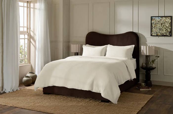 Penataan kamar tidur yang nyaman dengan tempat tidur mewah yang dilengkapi perlengkapan tidur putih, kepala tempat tidur berwarna gelap, dan meja samping tempat tidur dengan dekorasi. Jendela besar dengan tirai yang mengalir
