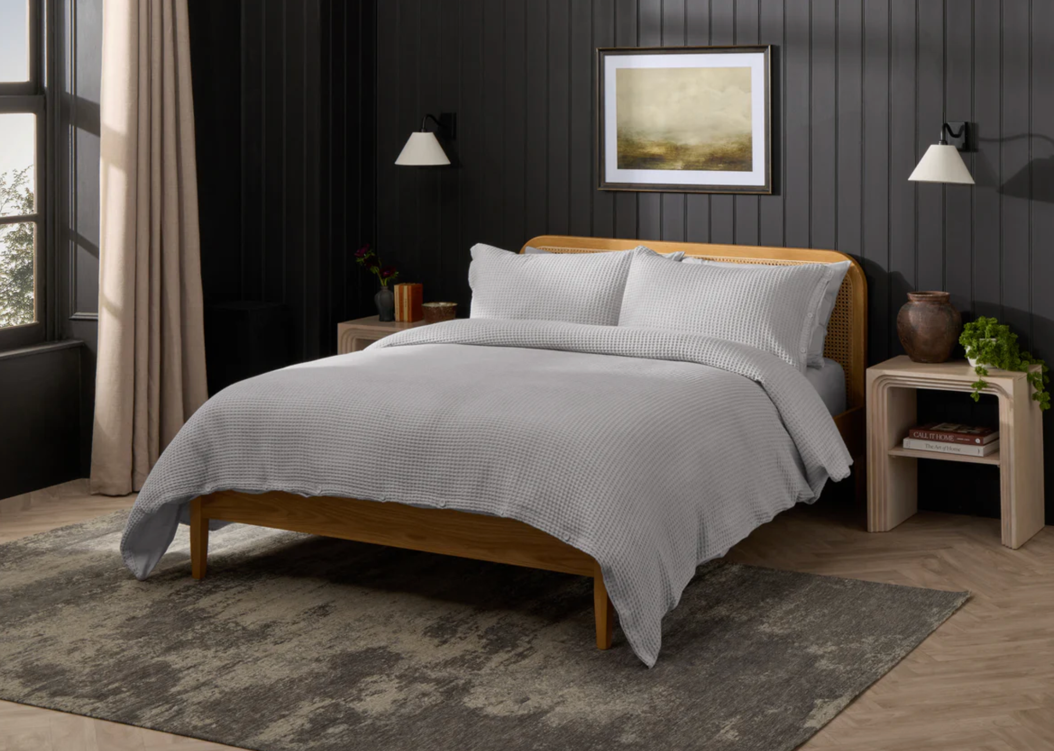 Kamar tidur modern dengan tempat tidur nyaman, perlengkapan tidur bergaris, bingkai kayu, dan meja samping tempat tidur. Lampu dinding dan seni berbingkai menciptakan suasana yang tenang dan mengundang