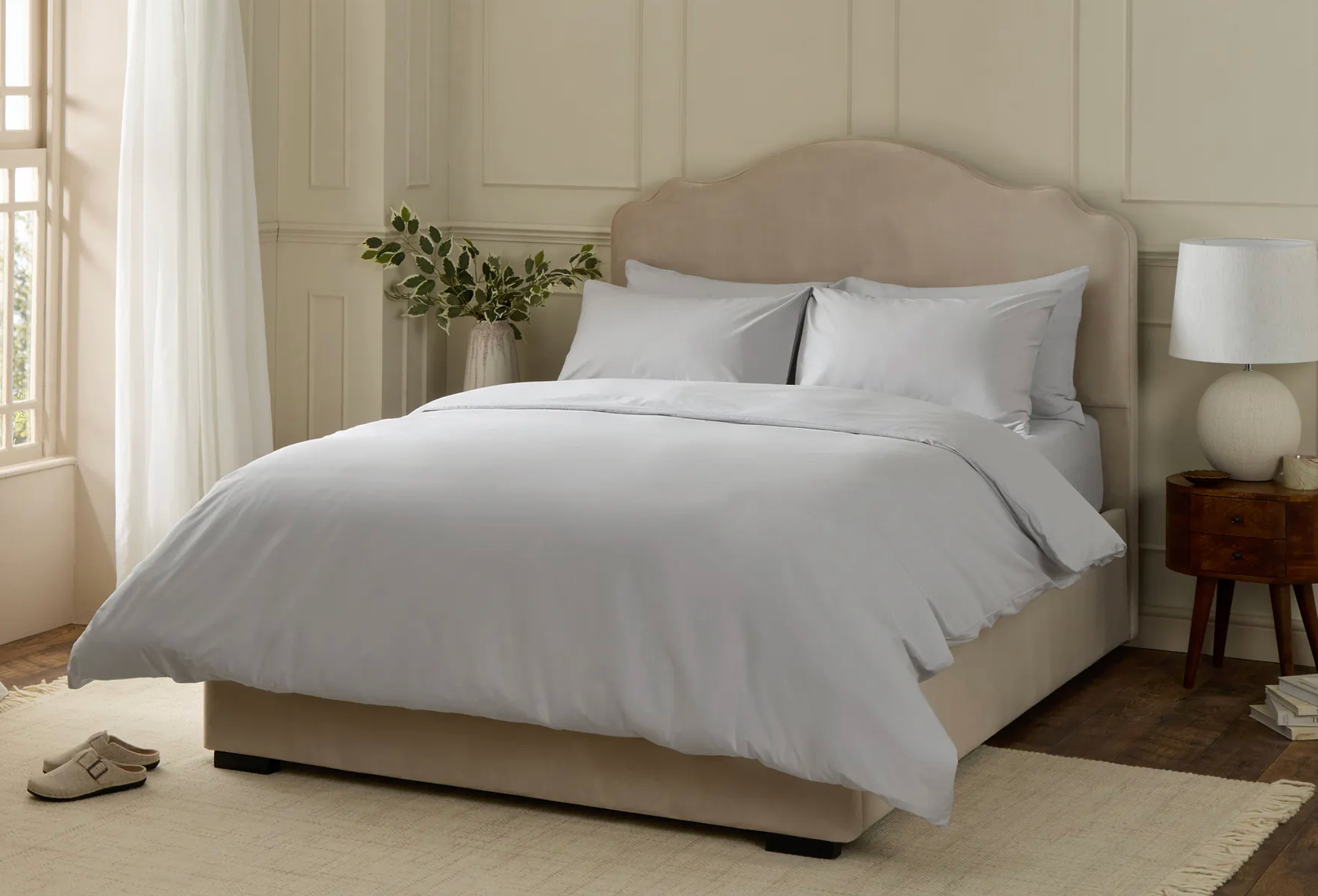 Tempat tidur yang tertata rapi dengan tempat tidur putih di kamar nyaman yang dilengkapi meja samping tempat tidur, lampu, dan tanaman, menunjukkan penataan kamar tidur yang nyaman