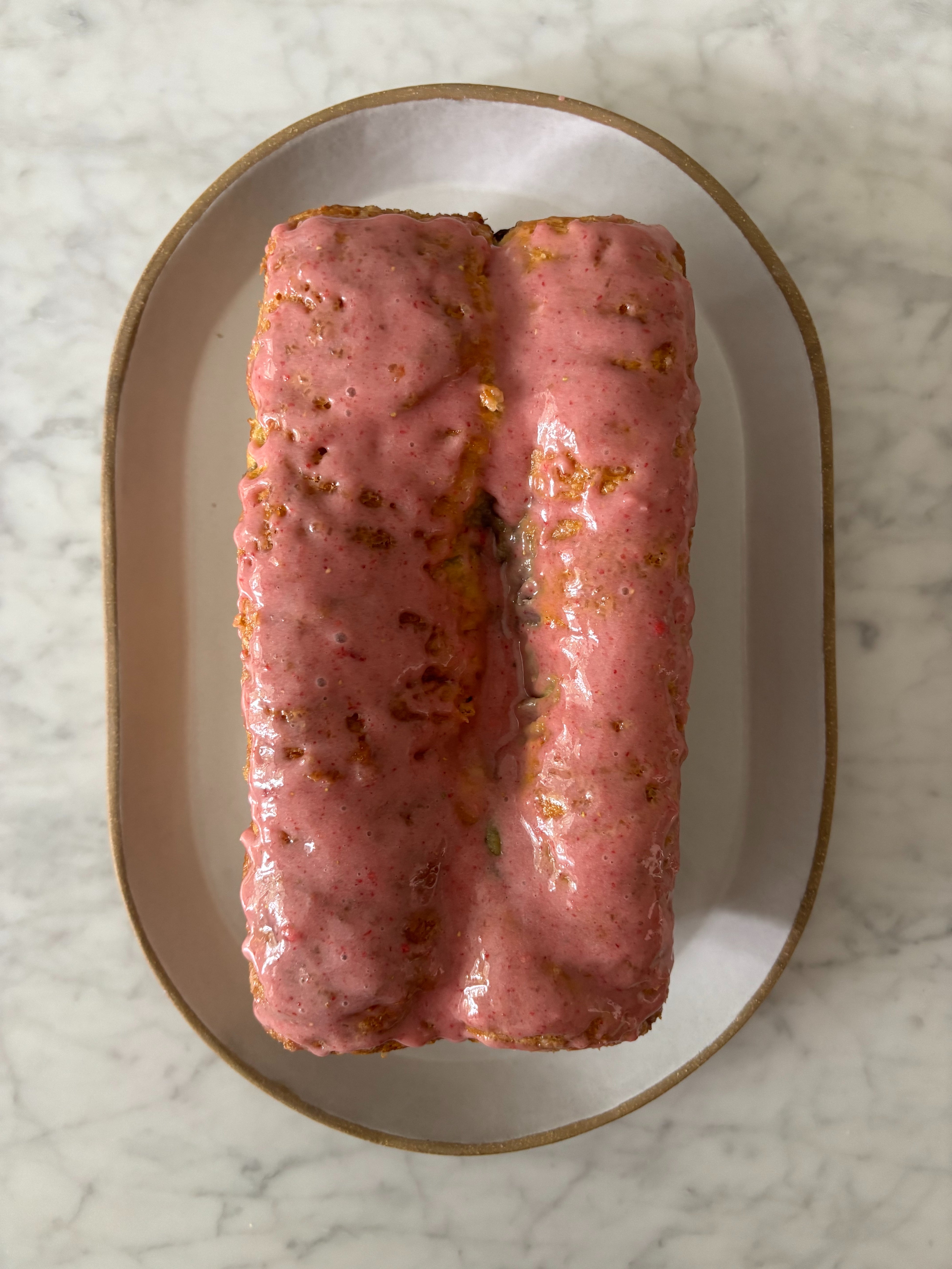 Loaf cake covered with a glossy pink glaze on a white oval plate, set on a marble surface