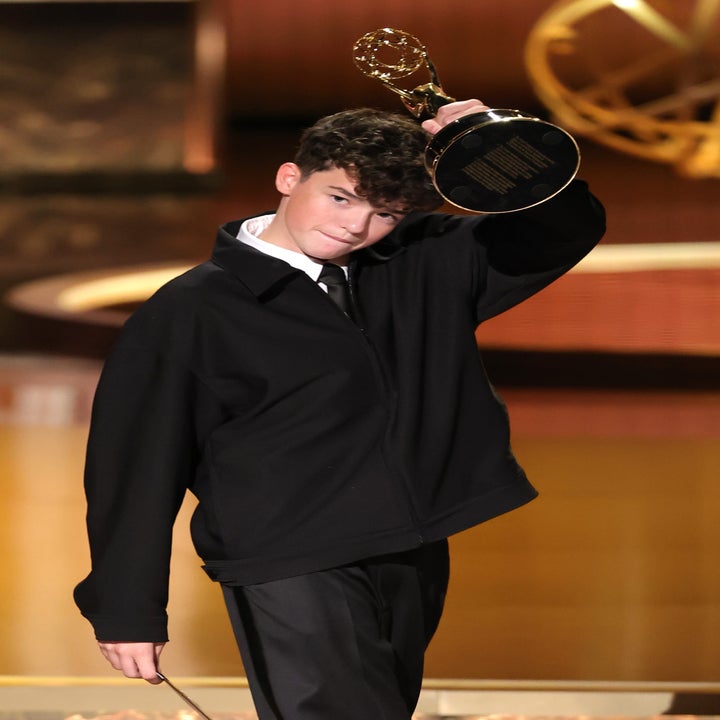 Person holding an award on stage, dressed in a loose-fitting black outfit with wide-legged pants. Stage has a large backdrop and decorative elements