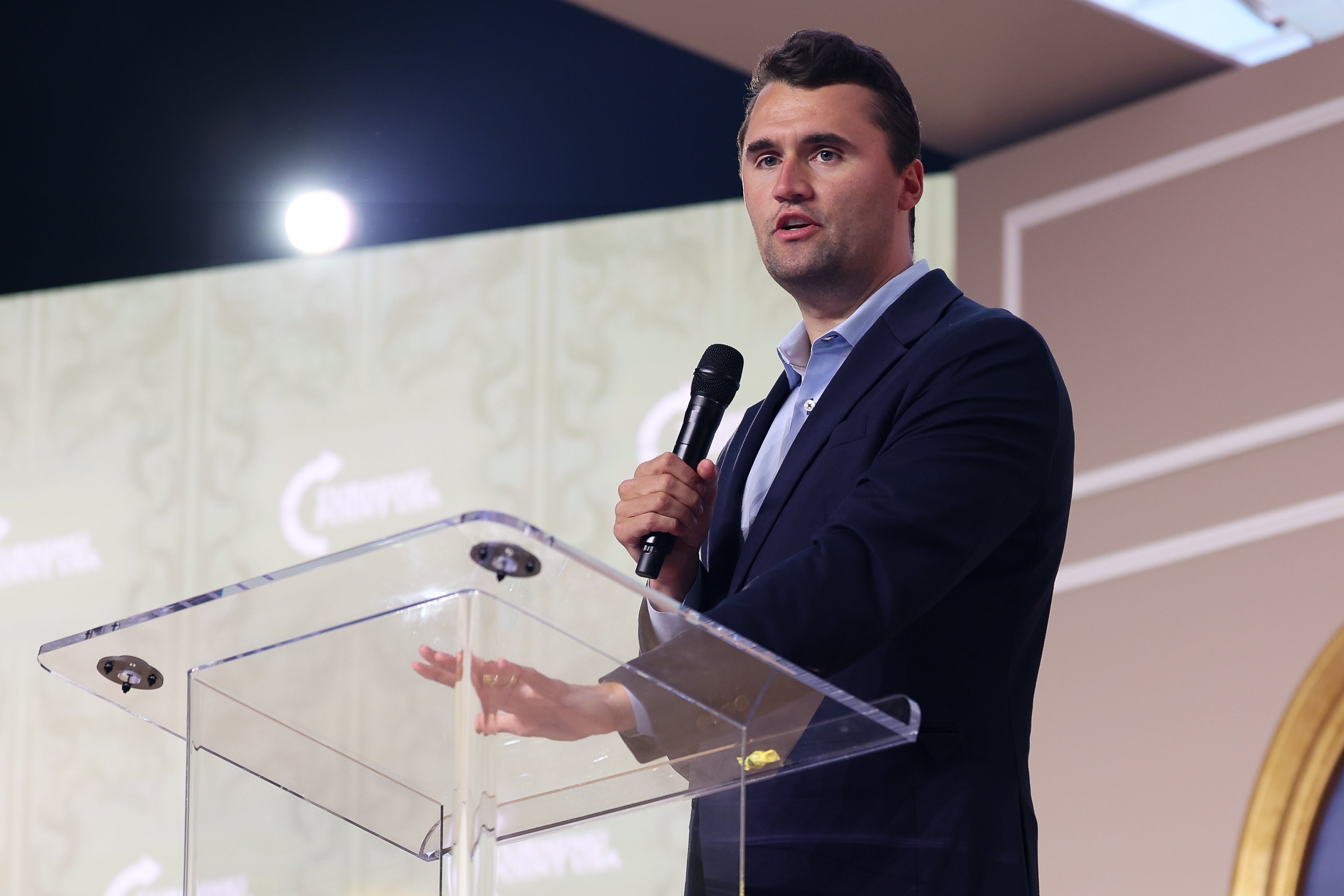 Charlie Kirk snakker ved et podium med en mikrofon, iført dressjakke og skjorte, mot en dekorativ bakgrunn