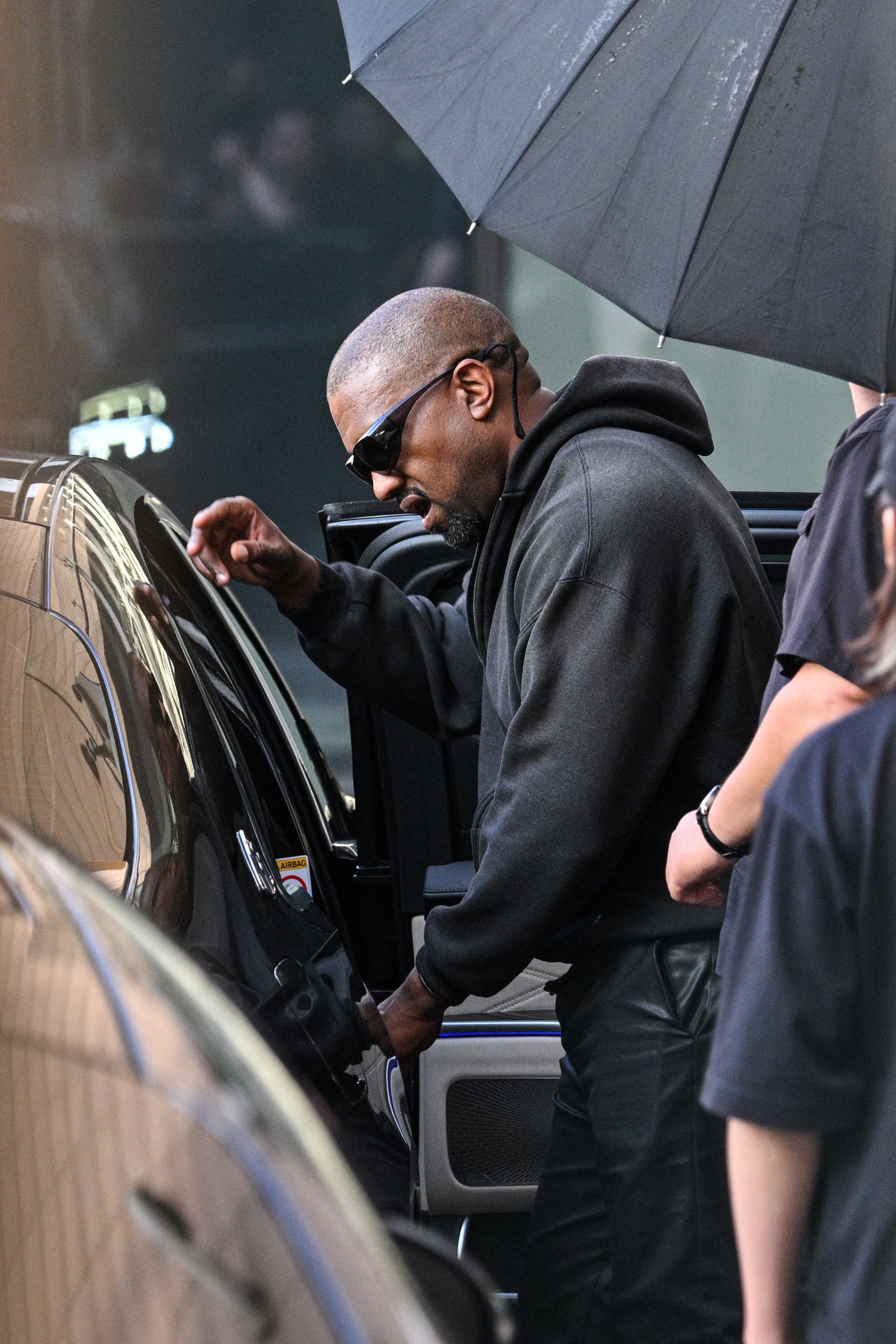 Kanye, iført hettegenser og solbriller, går ut av en bil, assistert av andre med en paraply over hodet
