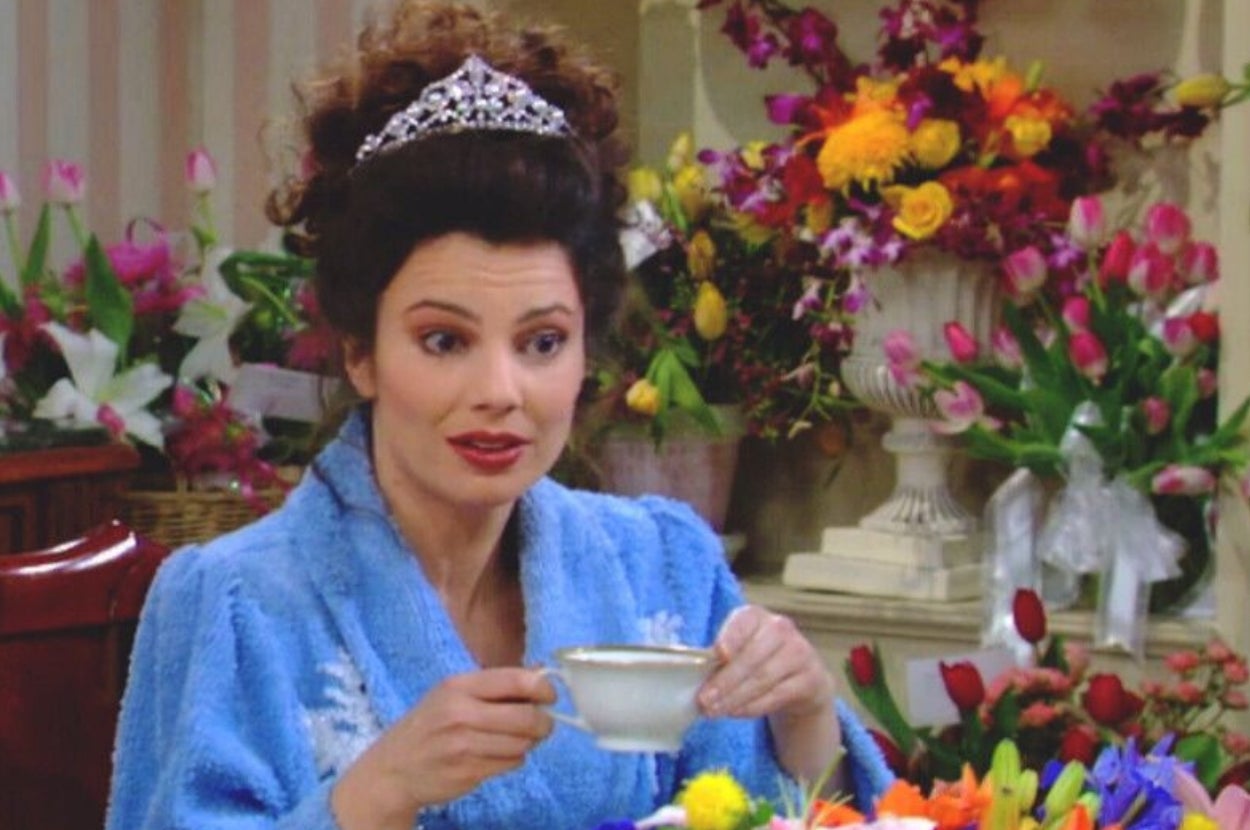 Person wearing a tiara and blue robe holds a cup, surrounded by colorful flowers, appearing surprised or engaged in conversation