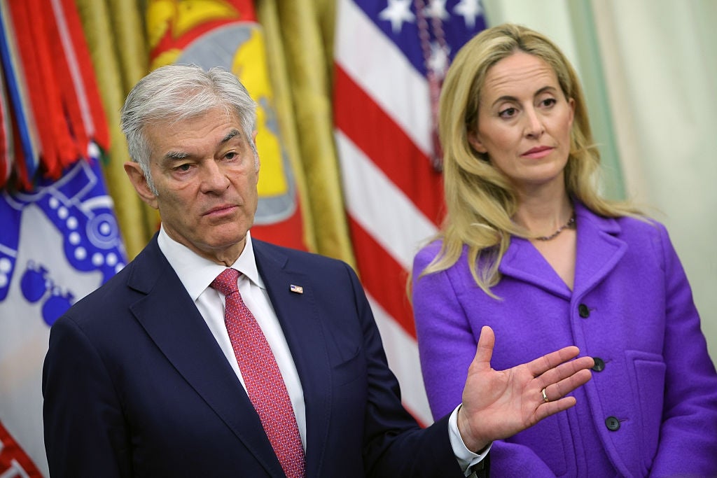 A antheral   successful  a suit   and necktie  speaks astatine  a property   event, portion    a pistillate   successful  a purple blazer stands beside him, successful  a country   with subject   flags successful  the background