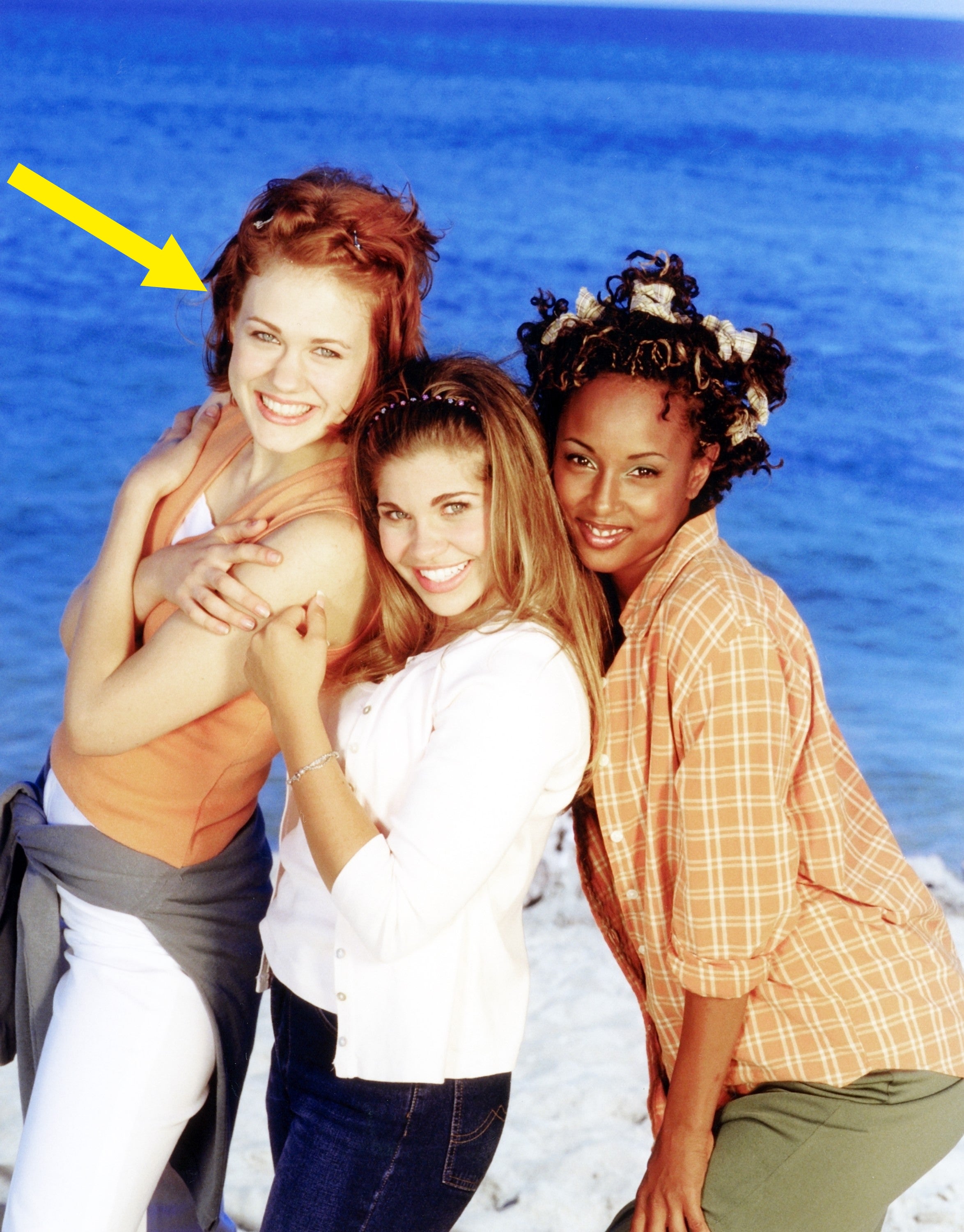 Three women airs  smiling connected  a formation  with the water  successful  the background. They deterioration  casual, beach-friendly clothing